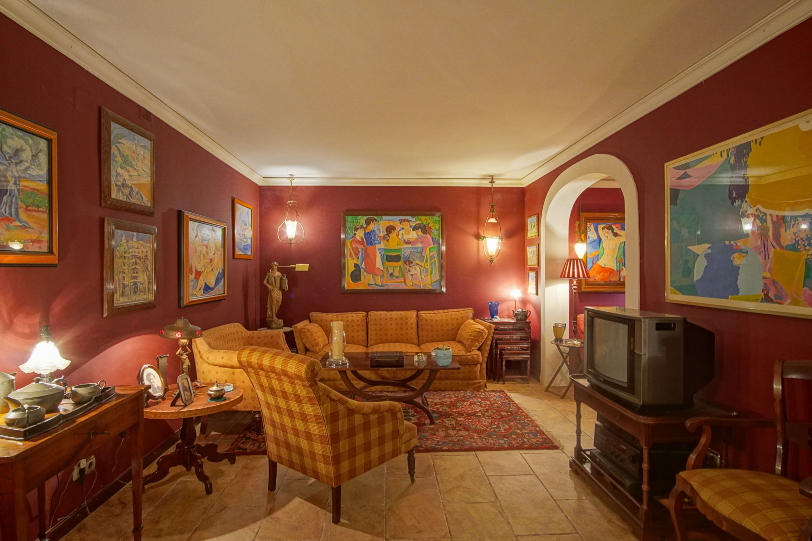 Wohnzimmer mit weinroten Wänden, rustikalem Fliesenboden und klassischen Polstermöbeln. Kunstwerke, warmes Licht und ein Rundbogen kennzeichnen diesen Raum.