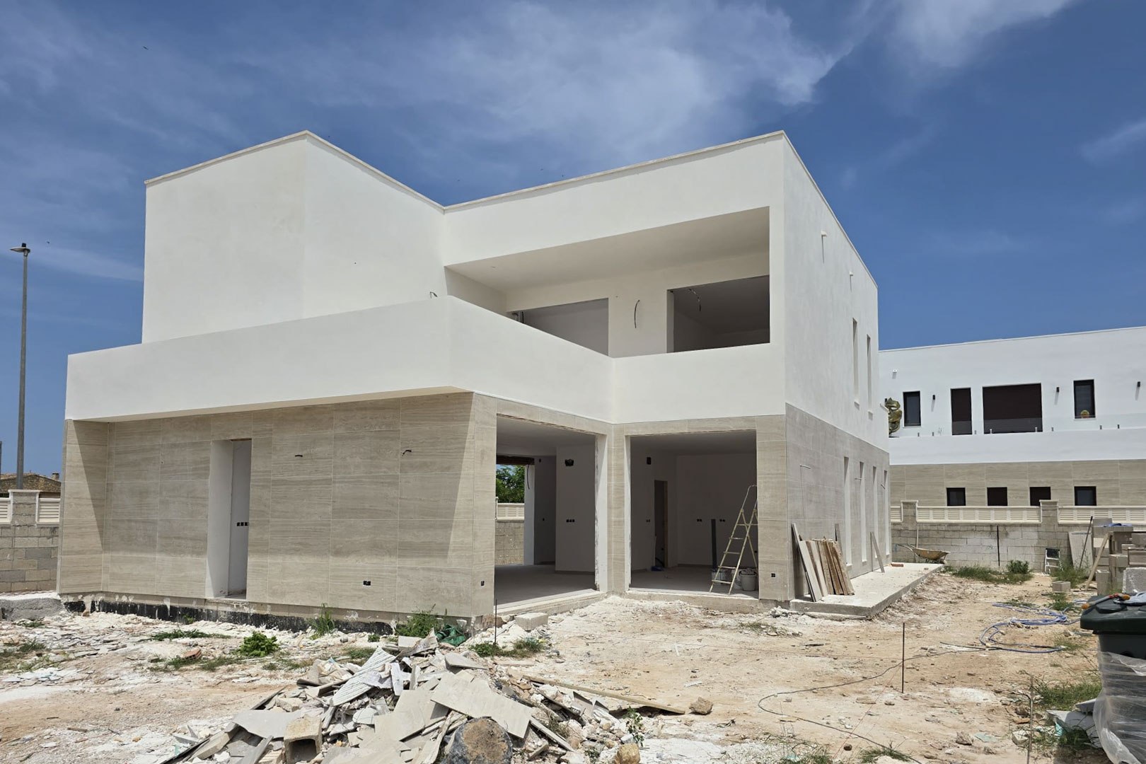 Außenansicht einer modernen Villa im Bau mit Natursteinverkleidung im Erdgeschoss, weißen geometrischen Formen und großen Öffnungen für bodentiefe Fensterfronten.