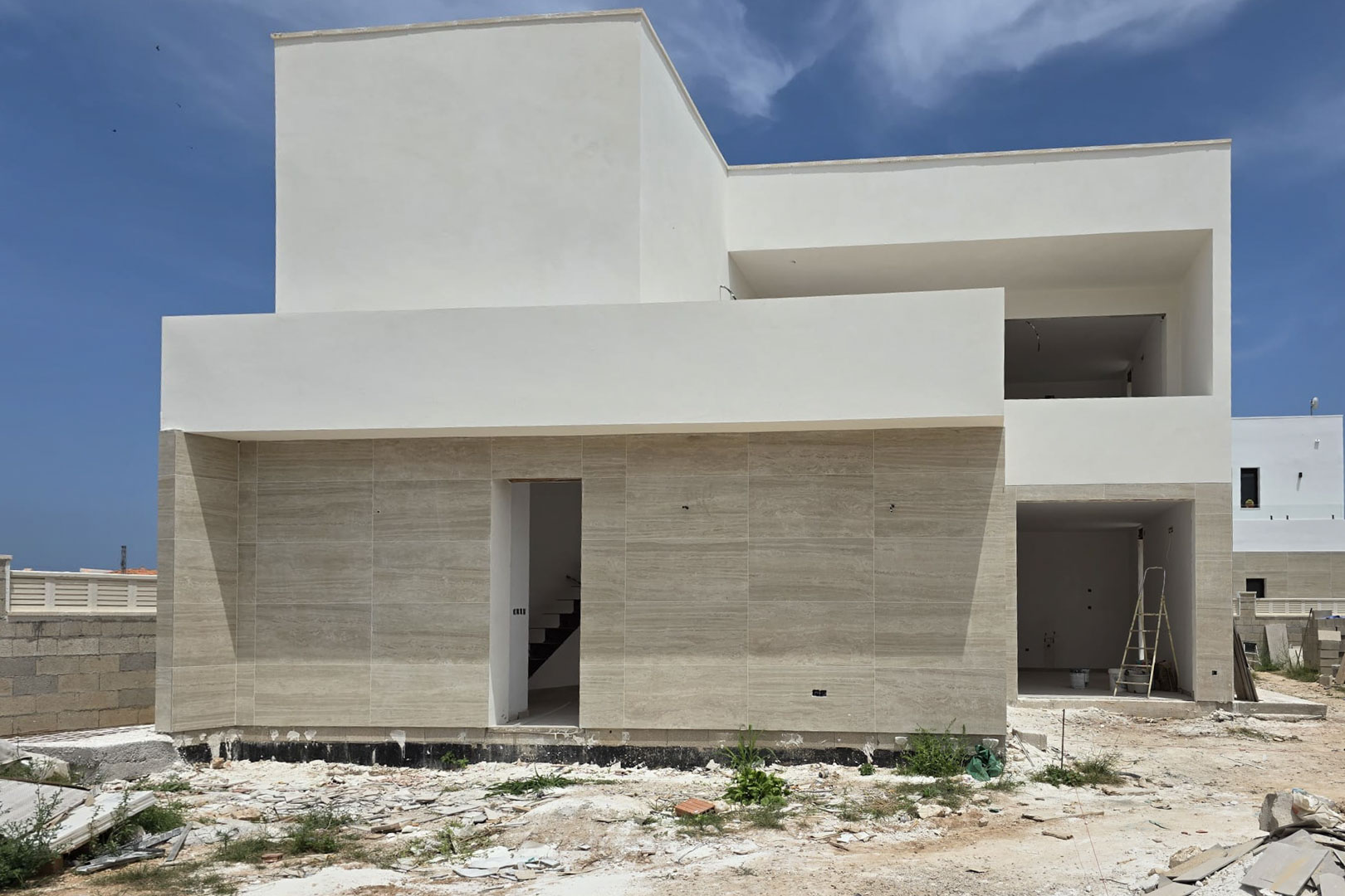 Außenfassade der im Bau befindlichen Villa in minimalistischem Design, mit heller Steinverkleidung im Erdgeschoss und weißen Oberflächen im Obergeschoss.