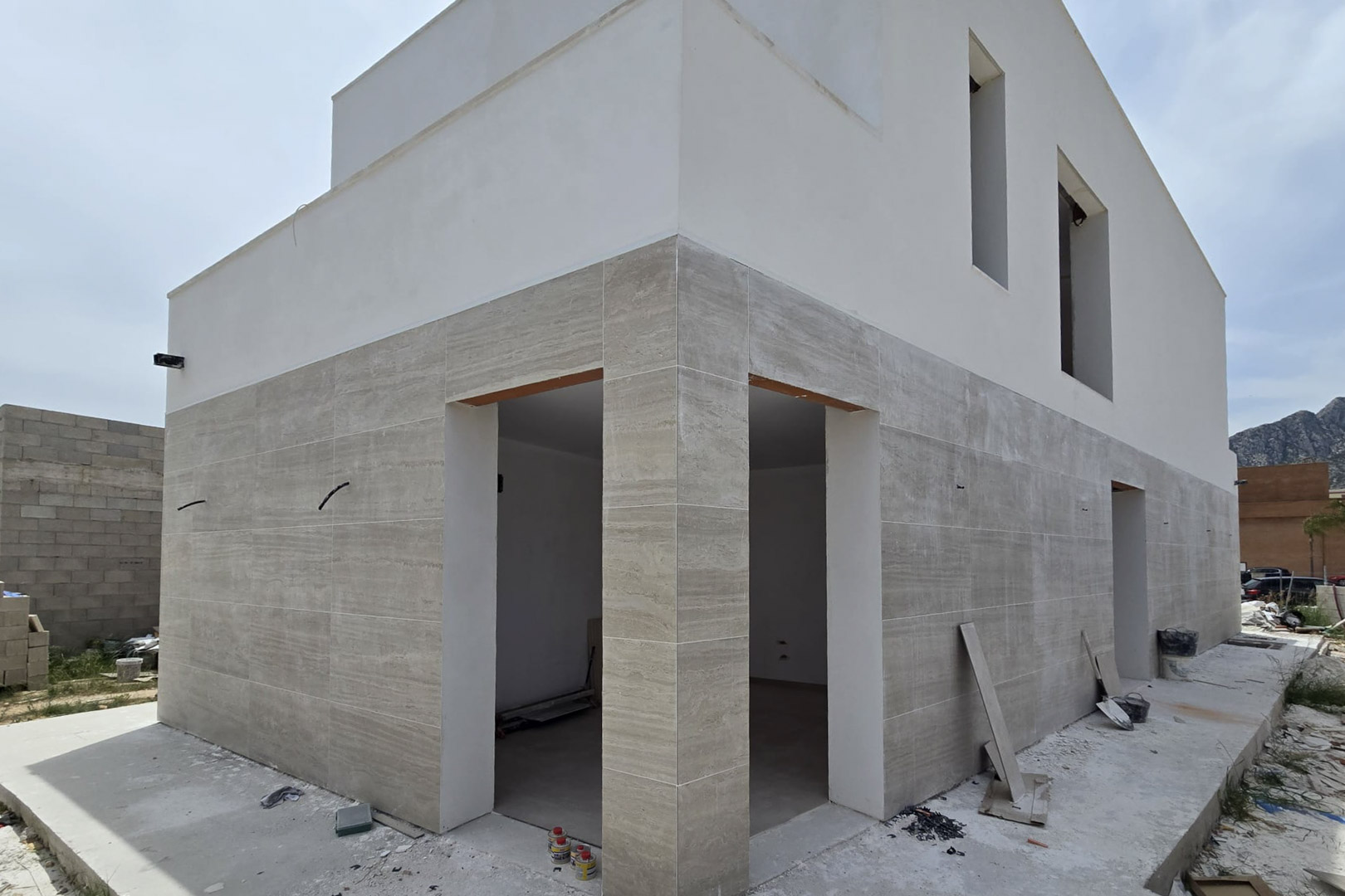 Außenfassade im Bau mit Steinverkleidung im Erdgeschoss und weißem Putz im Obergeschoss, die moderne geometrische Linien und große Fensteröffnungen zeigt.