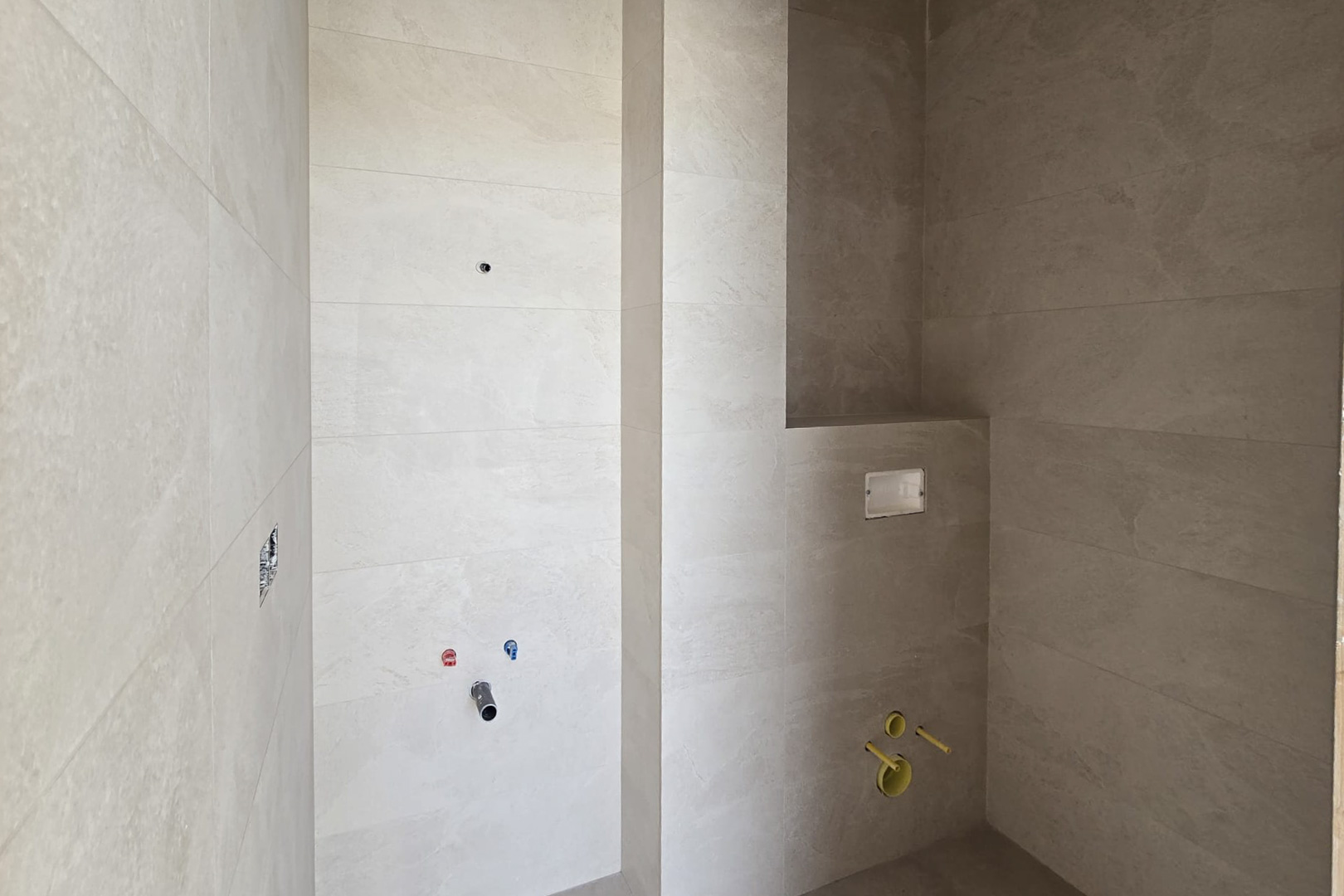 Badezimmer im Bau mit sandfarbenen Feinsteinzeugfliesen, Anschlüssen für ein Waschbecken und Vorbereitung für ein wandhängendes WC mit integrierter Nische.