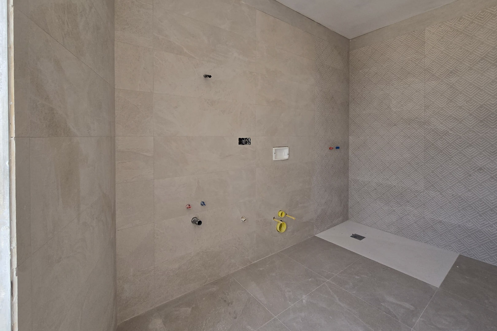 Badezimmer im Bau mit sandfarbenen Feinsteinzeugfliesen, geometrischer Akzentwand im Duschbereich und flacher weißer Duschwanne.