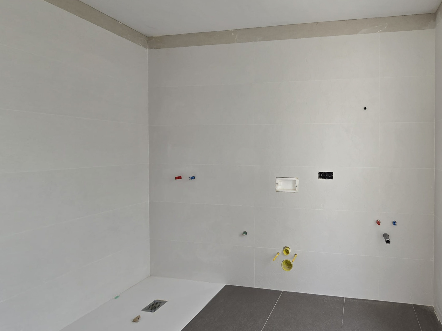 Badezimmer im Rohbau mit großformatigen weißen Fliesen, flacher Duschwanne und vorbereiteten Wasser- sowie Elektroanschlüssen für die Sanitärobjekte.