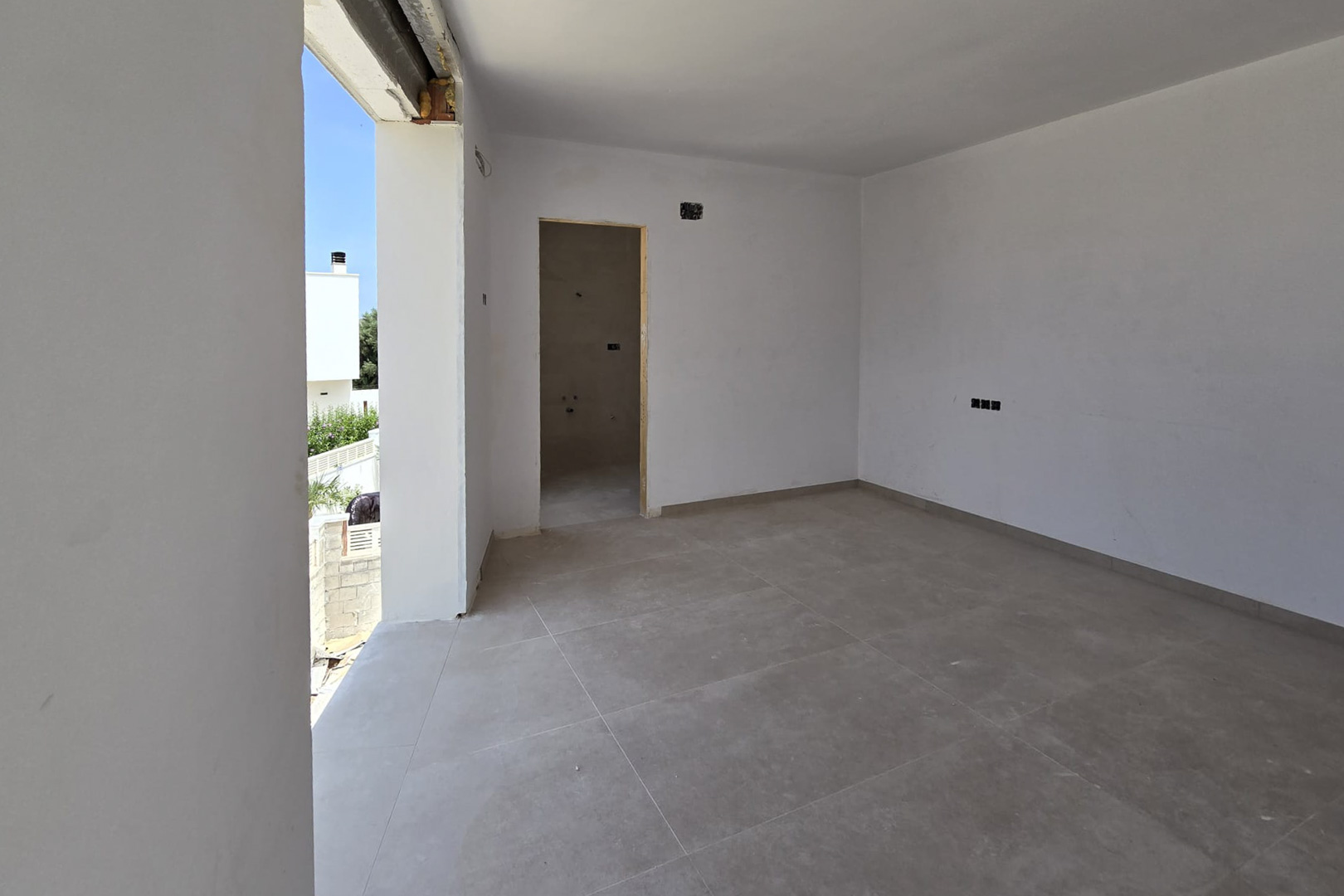 Innenraum im Bau mit großformatigen grauen Bodenfliesen, weißen Wänden und direktem Außenzugang. Der Eingang zu einem En-Suite-Badezimmer und Elektroanschlüsse sind sichtbar.