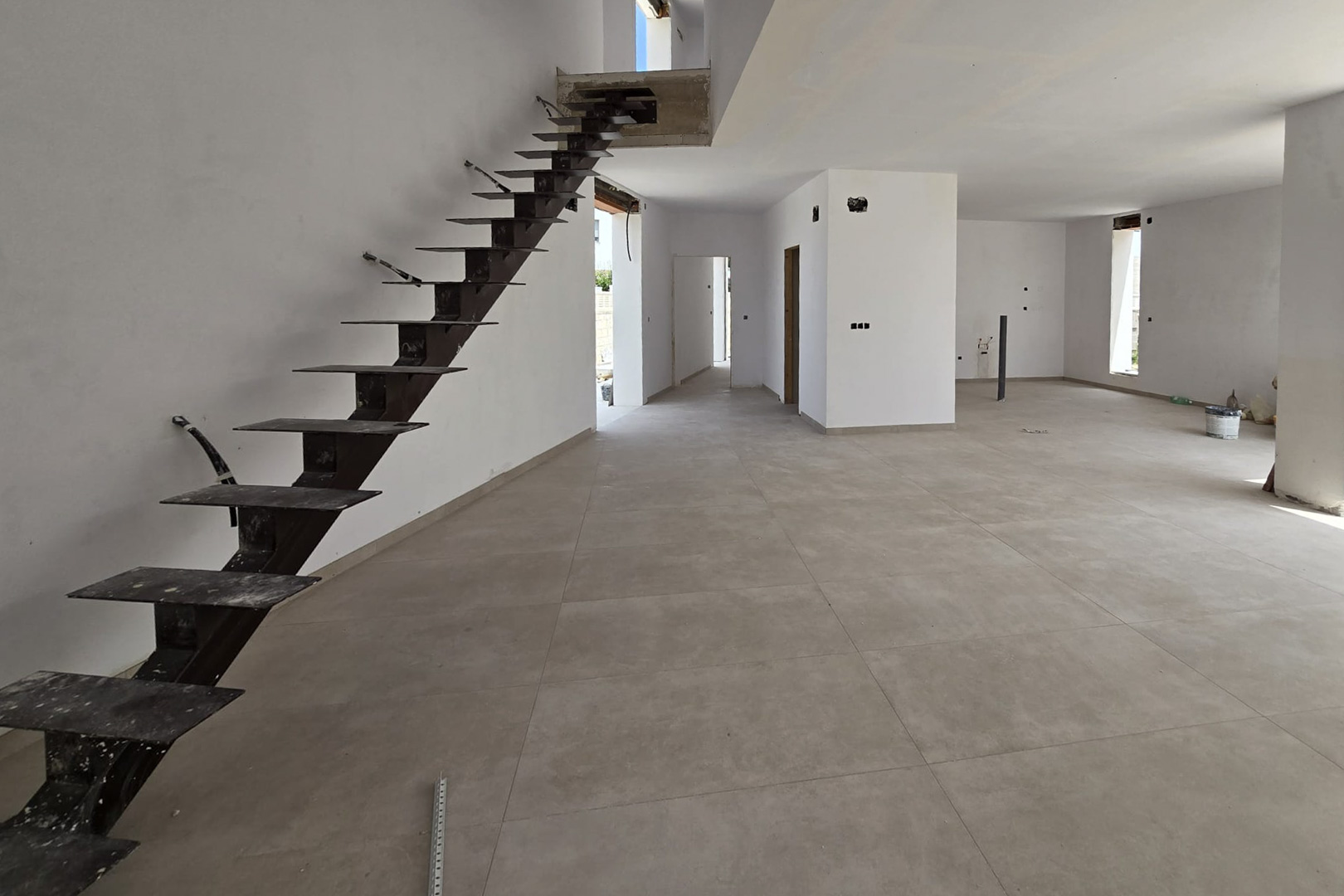 Offener Erdgeschossbereich im Bau mit großformatigen grauen Feinsteinzeugfliesen und einer freitragenden Metalltreppe zum Obergeschoss.