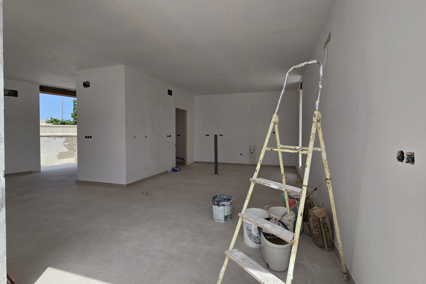 Offener Innenraum in der Endphase mit großformatigen Keramikfliesen und weißen Wänden. Sichtbar sind die Elektro-Vorinstallationen und der Zugang zum Außenbereich.