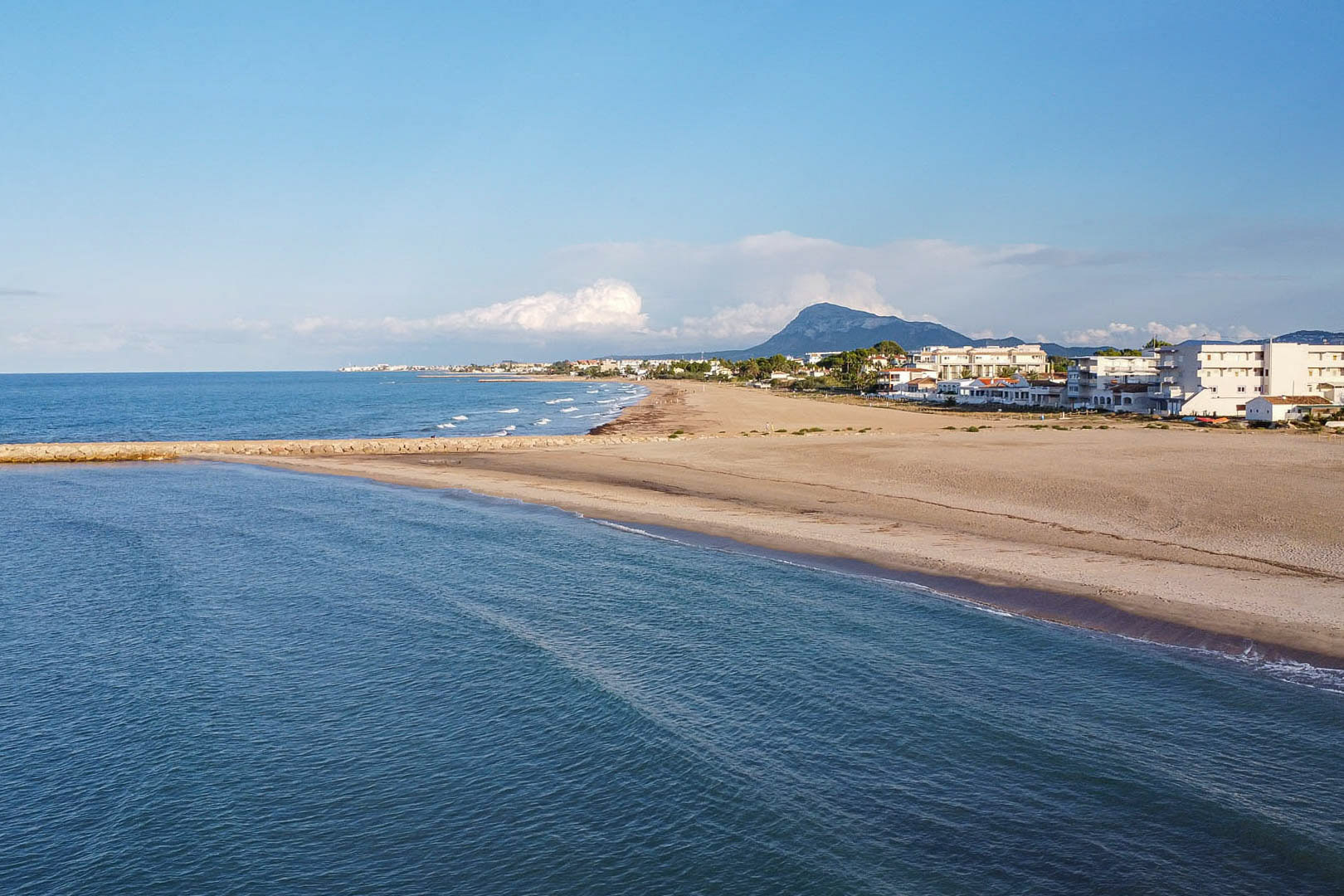 Panoramablick auf den weitläufigen Sandstrand in der Nähe, mit einer Steinmole, dem Mittelmeer und der Silhouette des Berges Montgó im Hintergrund.
