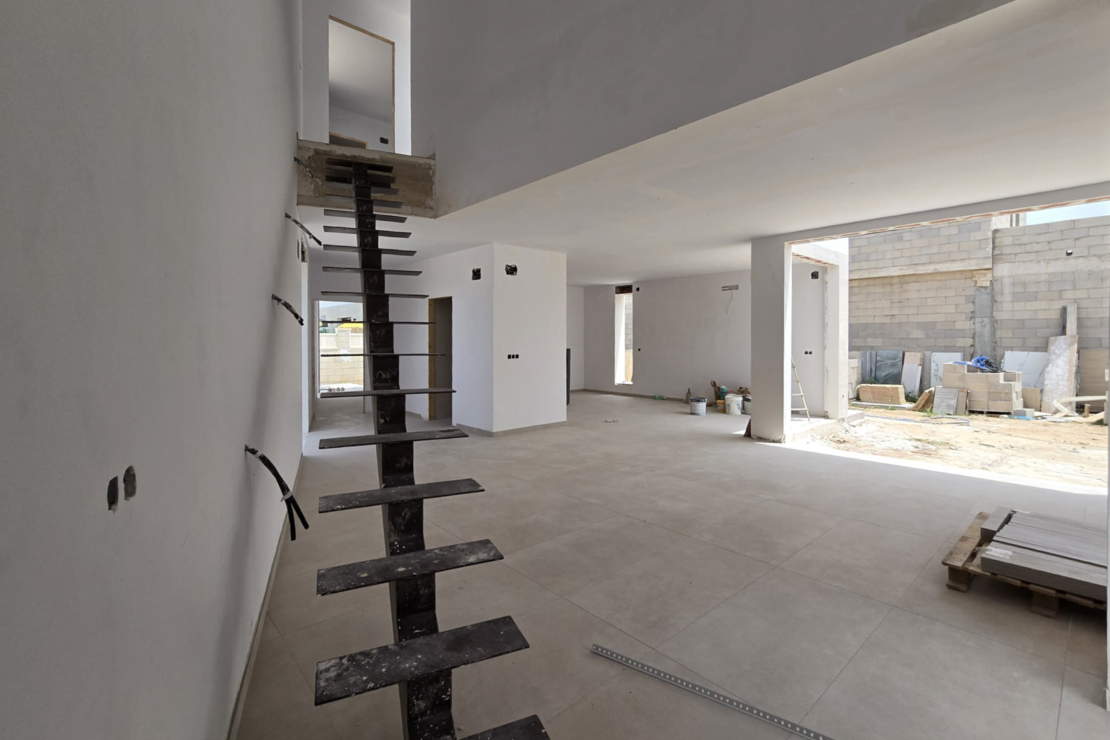 Rohbau des offenen Wohnbereichs mit einer modernen Metalltreppe, großformatigen Bodenfliesen und einem lichtdurchfluteten Luftraum über zwei Etagen.