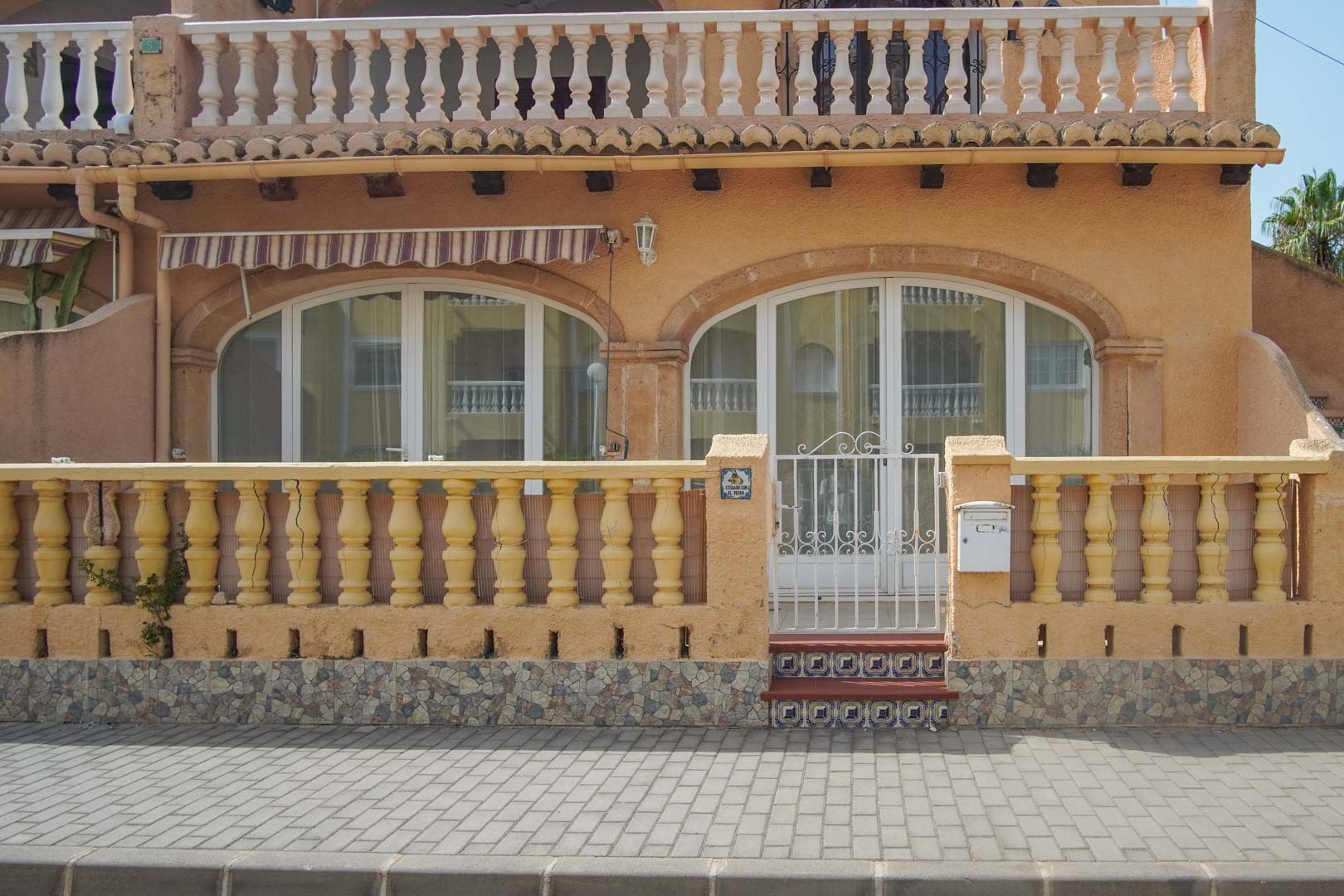 Außenansicht des Erdgeschosses mit klassischer Balustrade, weißem Metalltor und bogenförmigen Glastüren, die zur Terrasse führen.