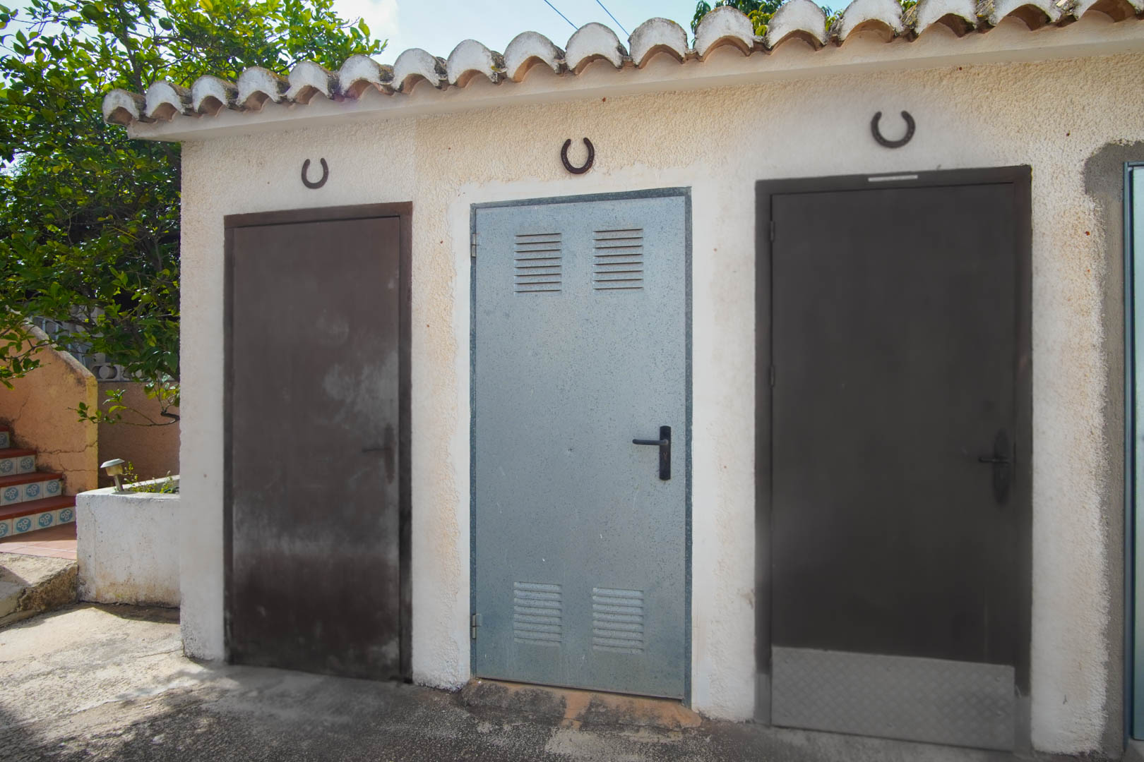 Außenlagerbereich mit drei Metalltüren unter einem traditionellen Ziegeldach, dekoriert mit Hufeisen an einer weiß verputzten Wand.