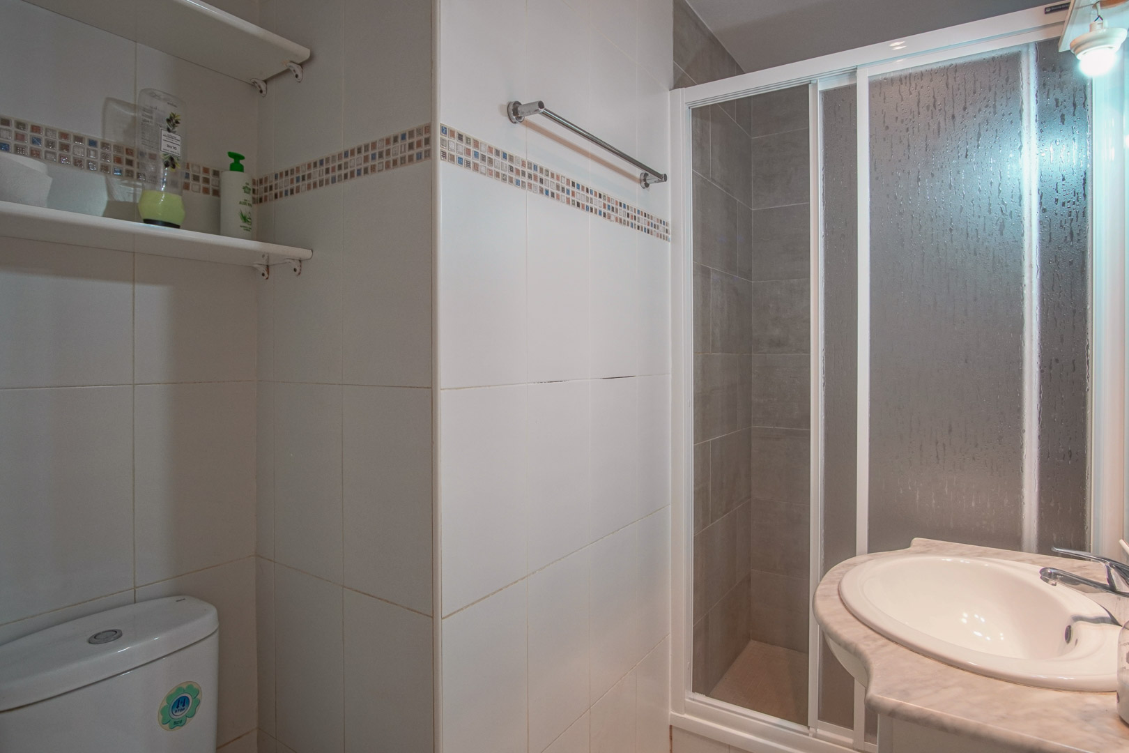 Badezimmer mit weiß gefliesten Wänden und Mosaikbordüre, ausgestattet mit einer Dusche mit Schiebetür und einem in eine Marmorplatte eingelassenen Waschbecken.