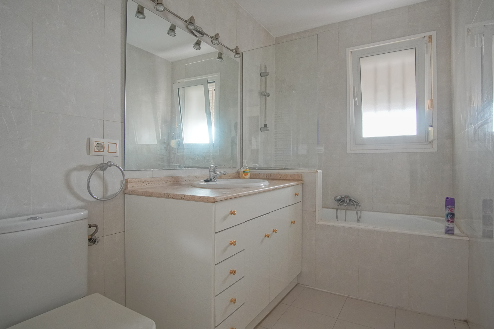 Badezimmer mit weißem Waschtisch, Marmorplatte und Badewanne mit Glasabtrennung. Ein Fenster sorgt für natürliches Licht und Belüftung im Raum.