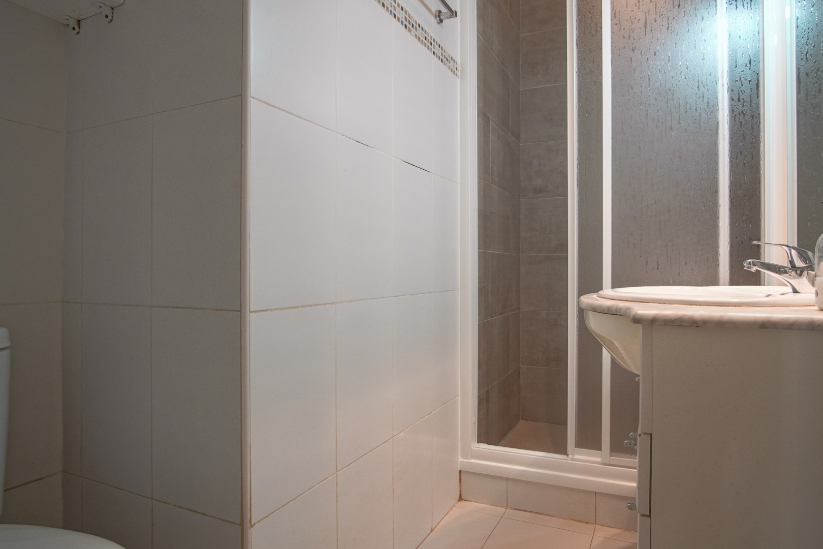 Funktionales Badezimmer mit weiß gefliesten Wänden, einer Duschkabine mit Sichtschutz und einem Waschbecken auf einer Marmorplatte.