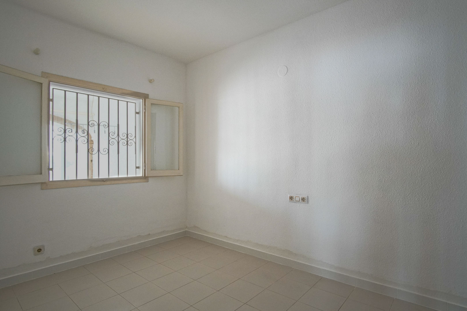 Helles Schlafzimmer mit weißen Strukturwänden, hellem Fliesenboden und Außenfenster mit dekorativen Sicherheitsgittern.