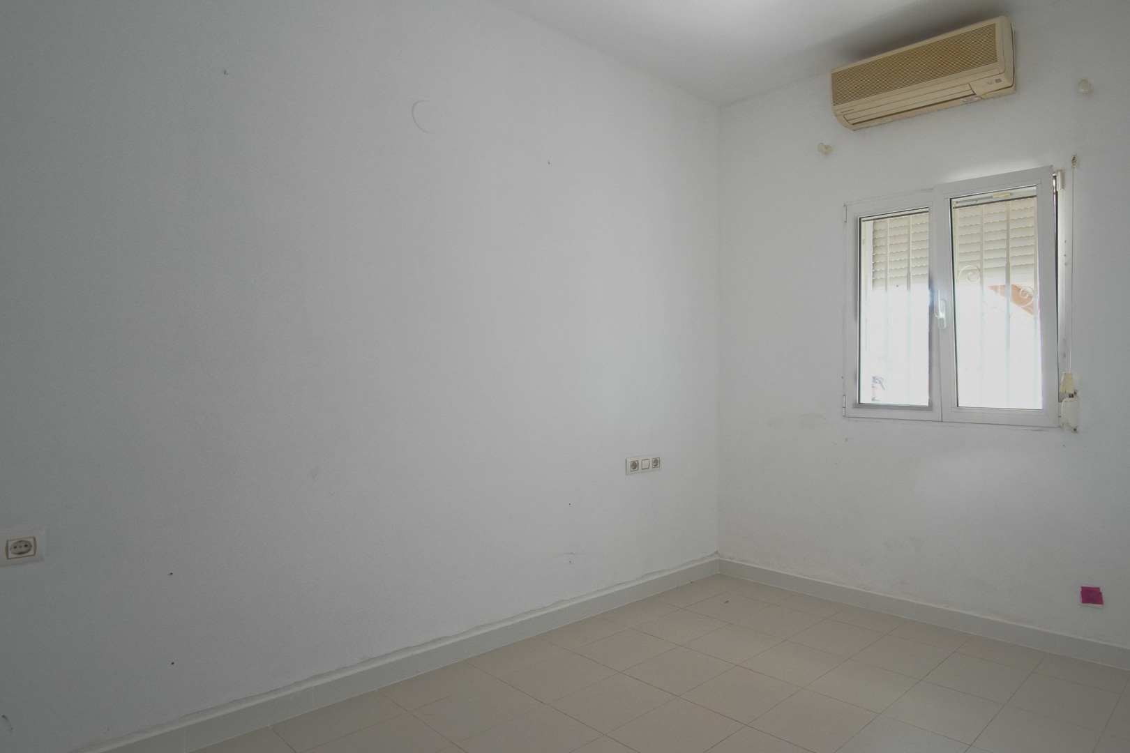 Helles Schlafzimmer mit weißen Wänden, Fliesenboden und Wandklimaanlage. Ausgestattet mit einem PVC-Fenster inklusive Rollladen und mehreren Steckdosen.