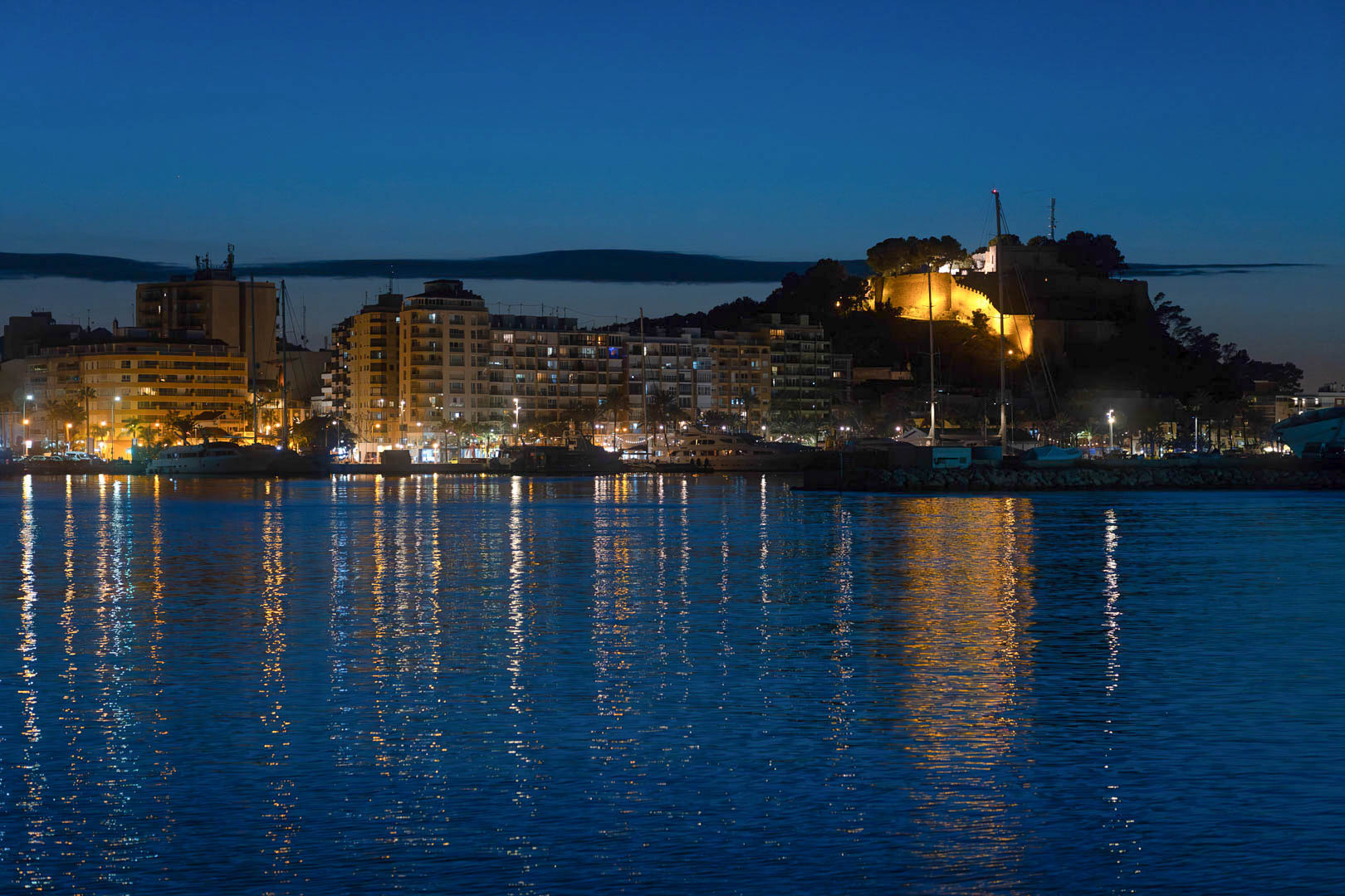 Nächtliche Ansicht des Hafens von Dénia mit der beleuchteten historischen Burg und der Stadtsilhouette, die sich im ruhigen Meer spiegelt.