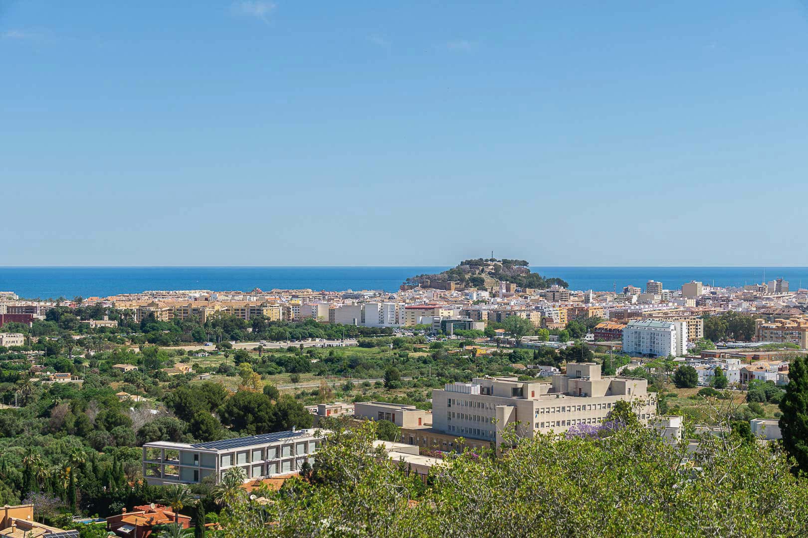 Panoramablick auf Dénia mit der historischen Burg auf dem Hügel, dem Mittelmeer im Hintergrund und der von Grünflächen umgebenen Stadtlandschaft.