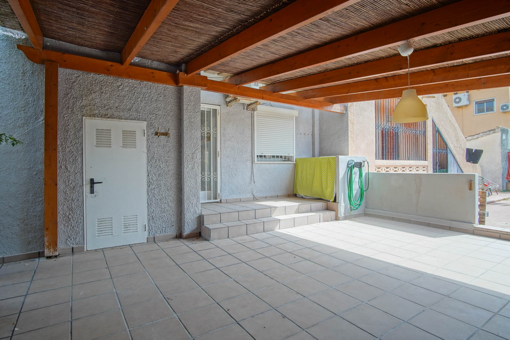 Überdachte Außenterrasse mit einer Holzpergola und Schilfmatten, Fliesenboden und direktem Zugang zum Wohnbereich über Stufen und eine weiße Sicherheitstür.