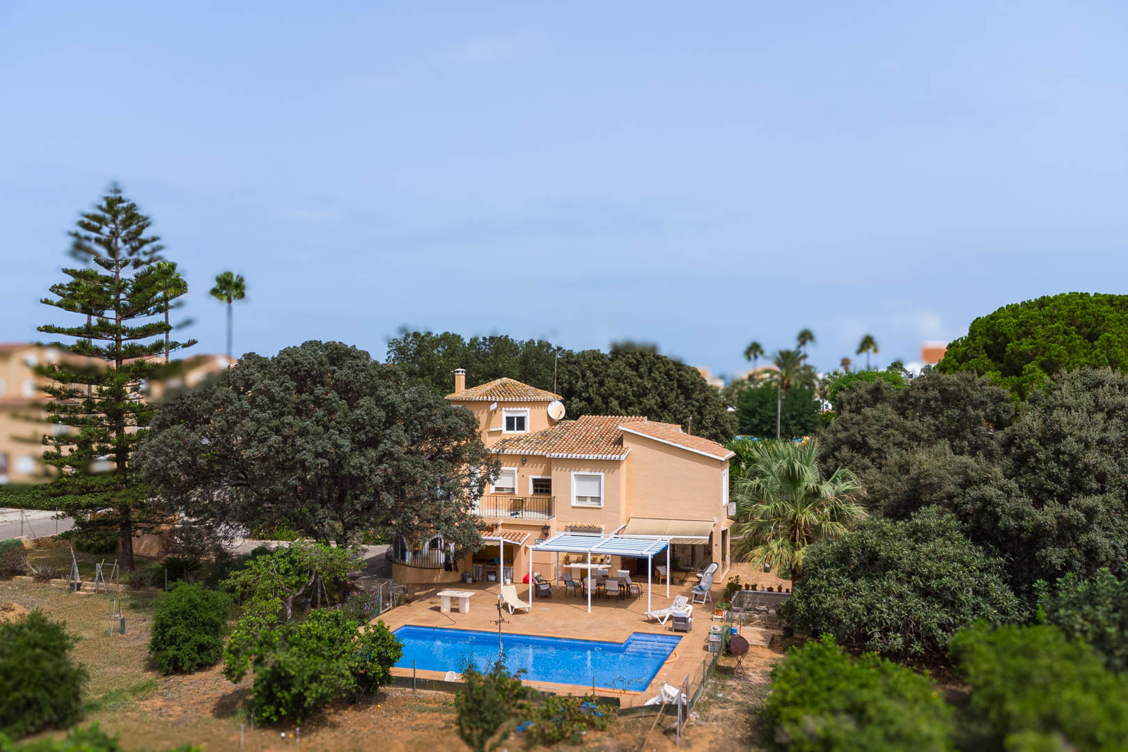 Außenansicht der Villa mit rechteckigem Pool, weißer Pergola-Terrasse und Essbereich im Freien, umgeben von üppiger mediterraner Vegetation und altem Baumbestand.