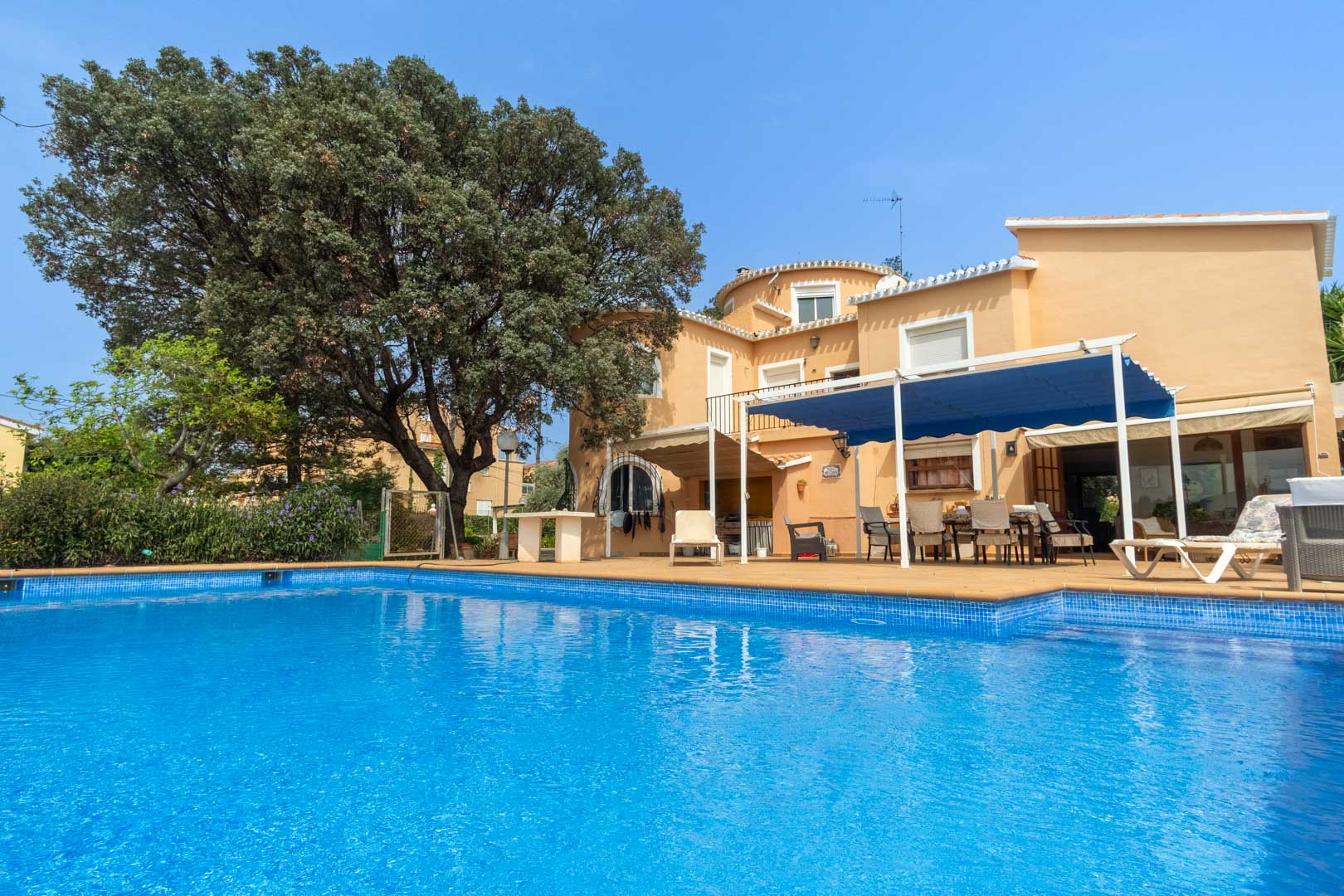Außenansicht des privaten Swimmingpools mit großer Sonnenterrasse und der mediterranen Villa mit einem durch eine blaue Markise beschatteten Essbereich.