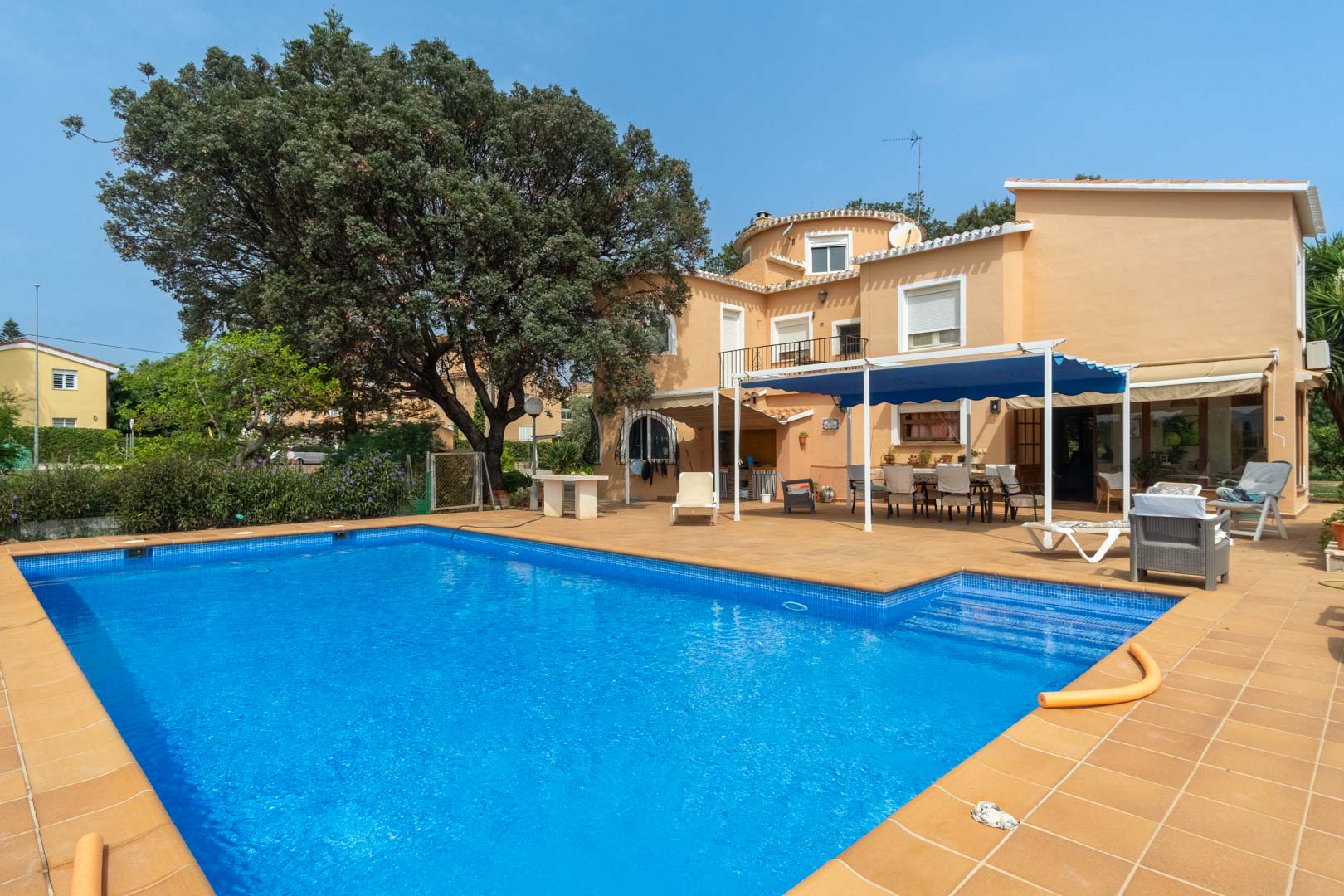 Außenansicht des rechteckigen Swimmingpools mit Terrakotta-Umrandung, mediterraner Fassade und einem schattigen Essbereich unter einer blauen Markise.
