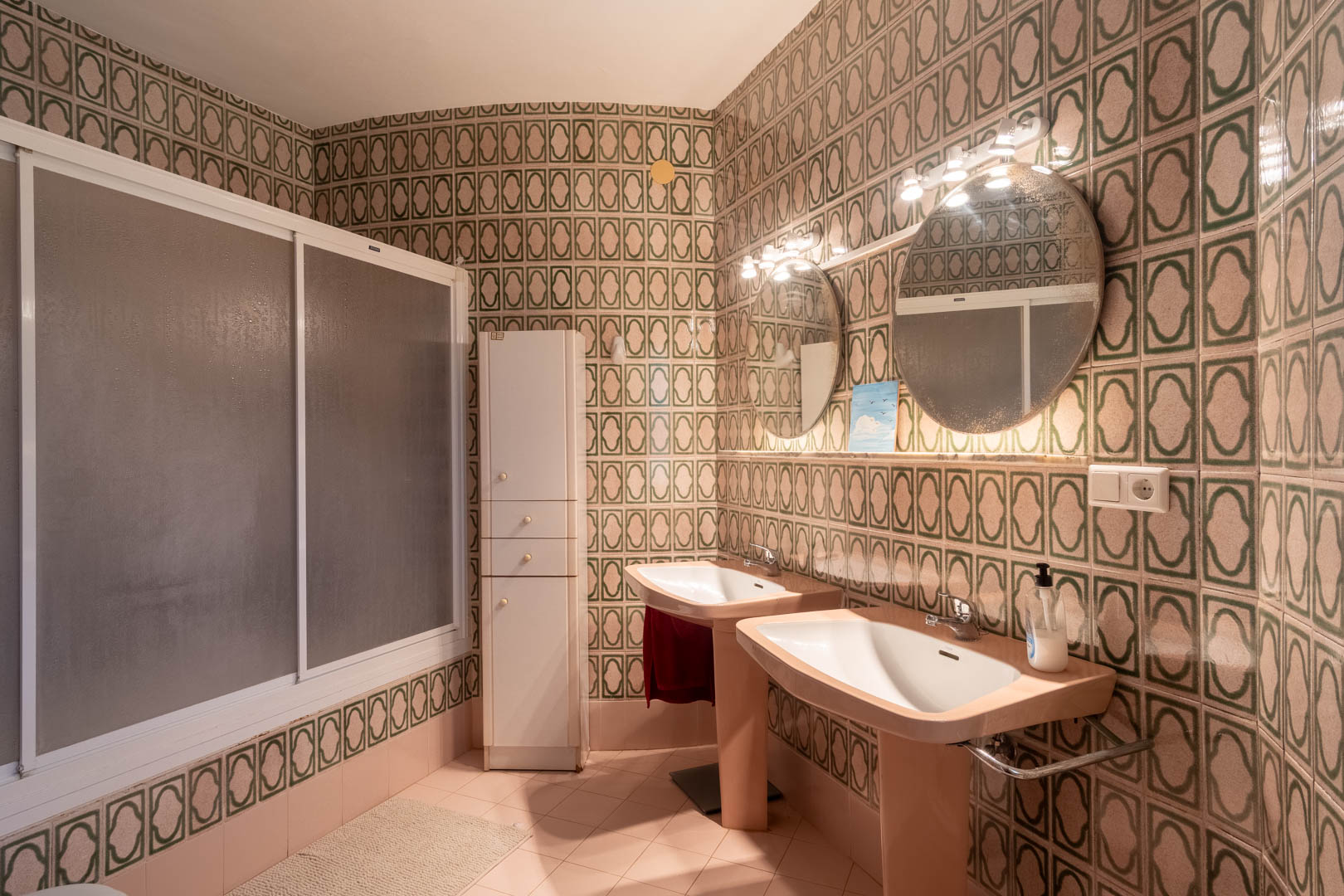Badezimmer im Retro-Stil mit Doppelwaschbecken, beleuchteten Rundspiegeln und grün gemusterten Wandfliesen. Mit begehbarer Dusche und weißem Hochschrank.