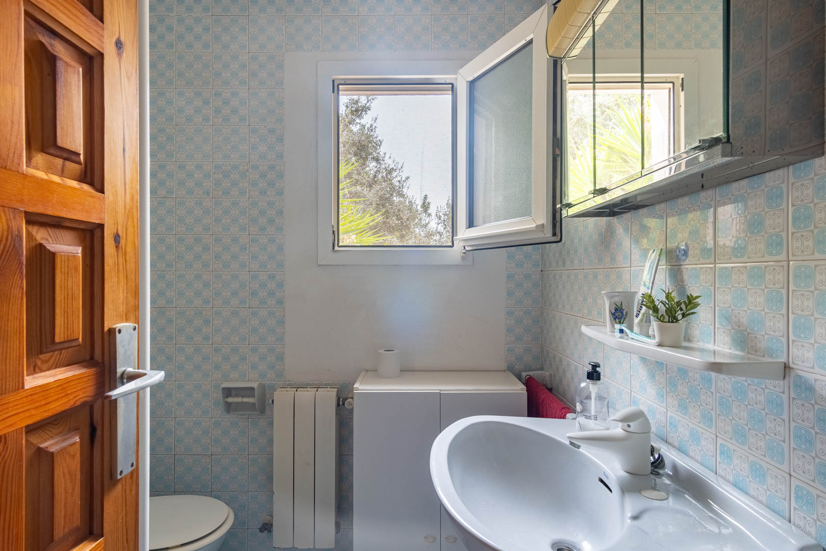 Badezimmer mit blau gemusterten Wandfliesen, weißem Waschbecken und einem Fenster mit Fliegengitter für natürliches Licht und Belüftung.