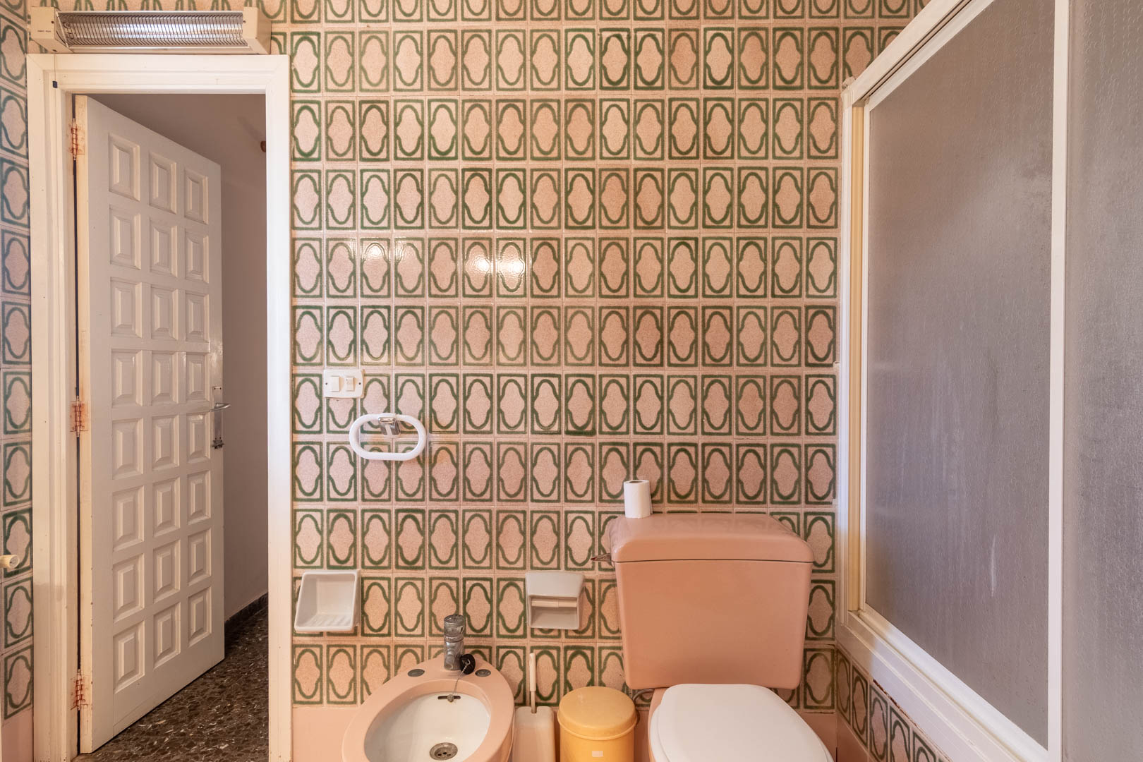 Badezimmer mit grün gemusterten Wandfliesen, rosa Porzellan-Sanitäranlagen inklusive Bidet und WC sowie einer weißen Paneeltür.