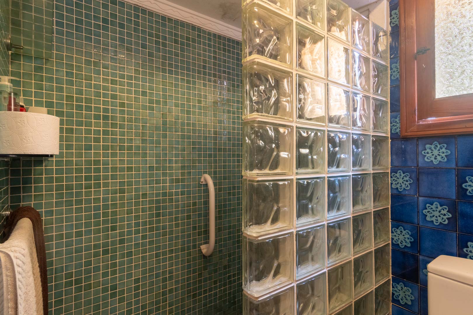 Badezimmer mit grünem Mosaik-Duschbereich und einer Trennwand aus Glasbausteinen. Dekorative blaue Fliesen und ein Holzfenster ergänzen die funktionale Gestaltung.