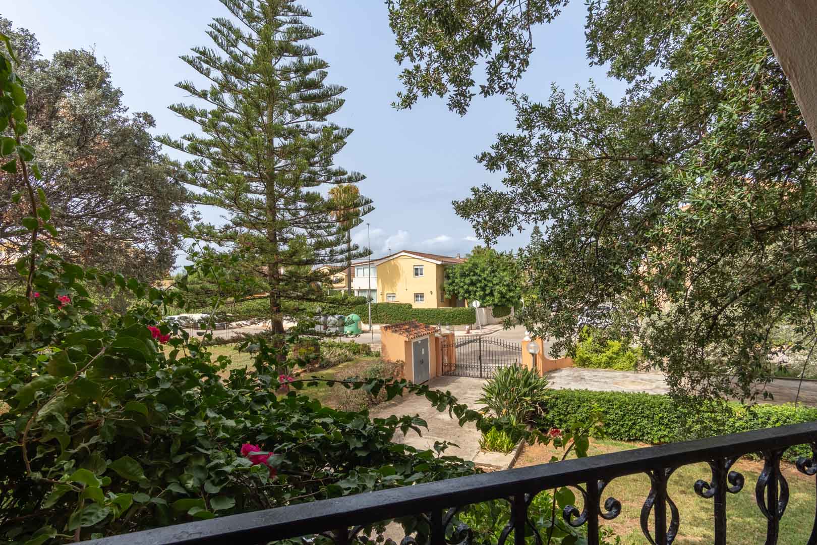 Blick von der oberen Terrasse mit Schmiedeeisengeländer auf den mediterranen Garten mit Pinien und Bougainvillea sowie das private Einfahrtstor.