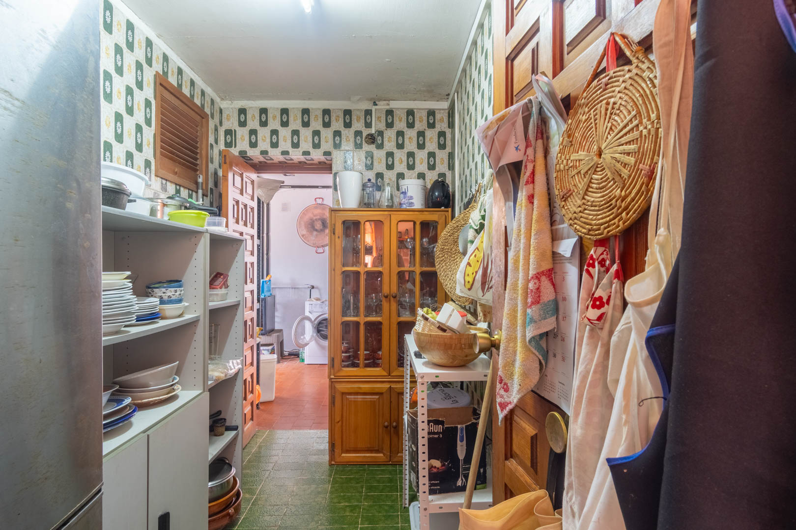 Funktionale Speisekammer mit Vintage-Fliesen, offenen Regalen für Geschirr und einer Holzvitrine, die direkt zum Waschraum führt.