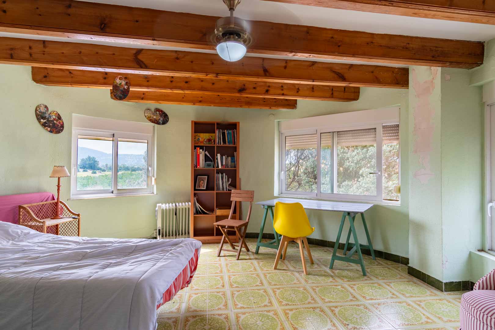 Helles Schlafzimmer mit sichtbaren Holzbalken, gemusterten Keramikfliesen und Arbeitsbereich. Große Fenster bieten einen weiten Blick auf die umliegende mediterrane Landschaft.