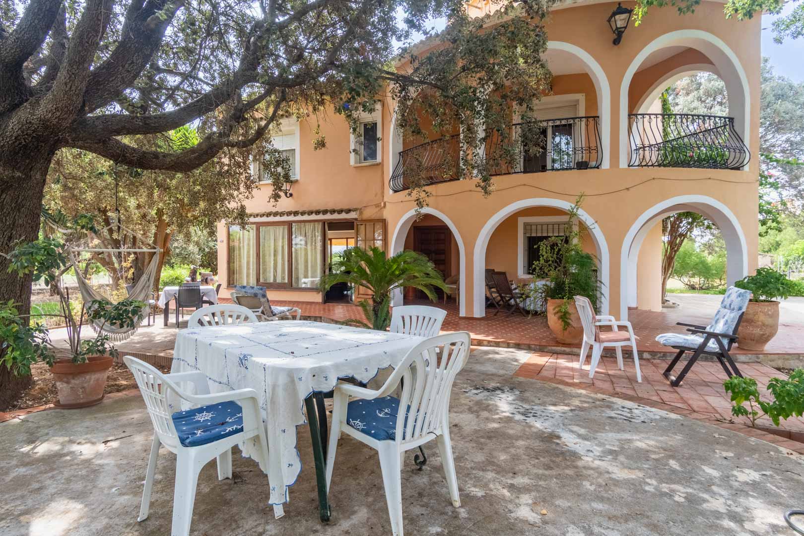 Mediterrane Fassade mit weißen Rundbogen-Veranden, von alten Bäumen beschatteten Terrassen und Essbereichen im Freien auf Terrakottaboden.