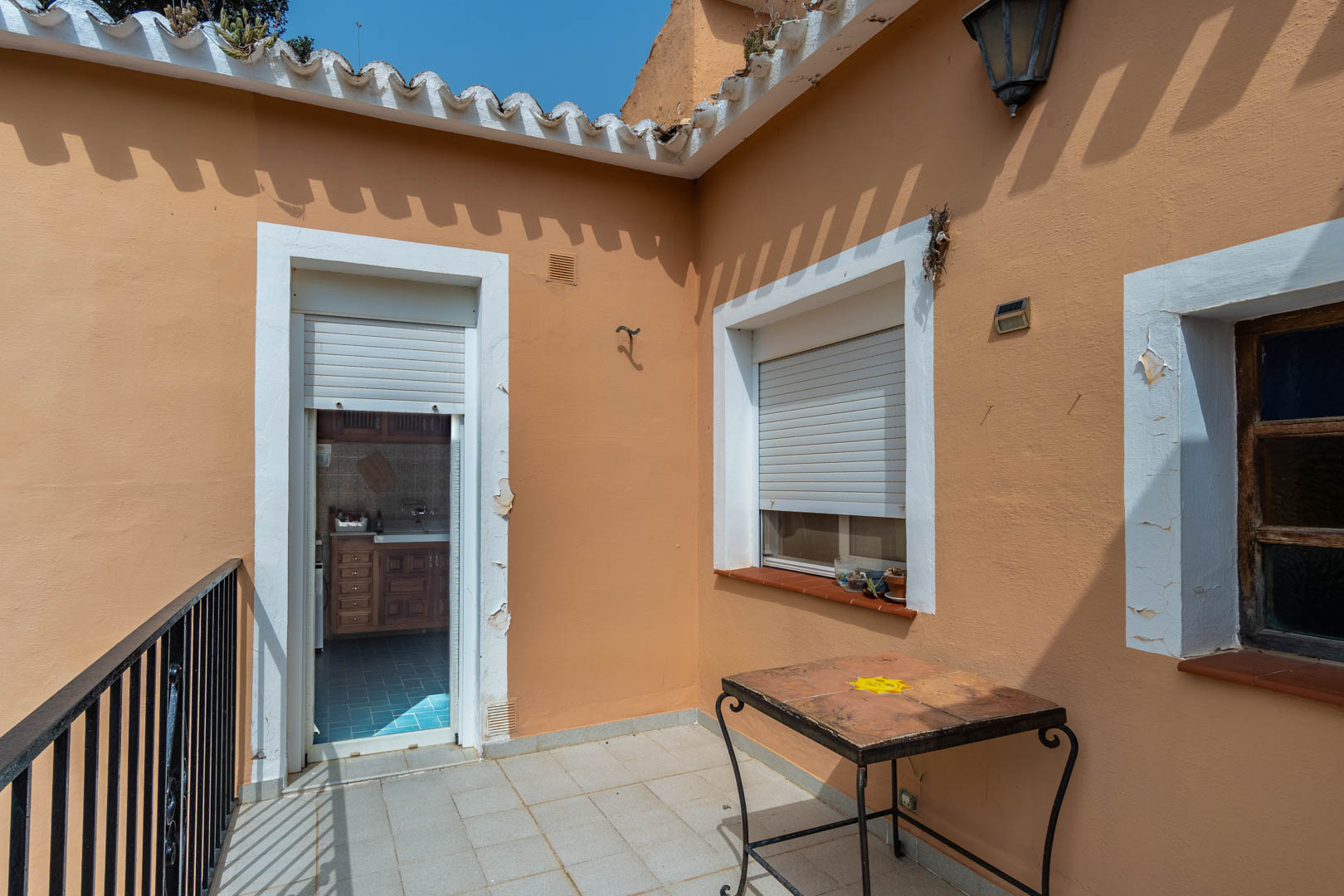 Obere Terrasse mit Fliesenboden und Schmiedeeisengeländer, die durch eine Tür mit Rollladen direkt mit der separaten Küche verbunden ist.