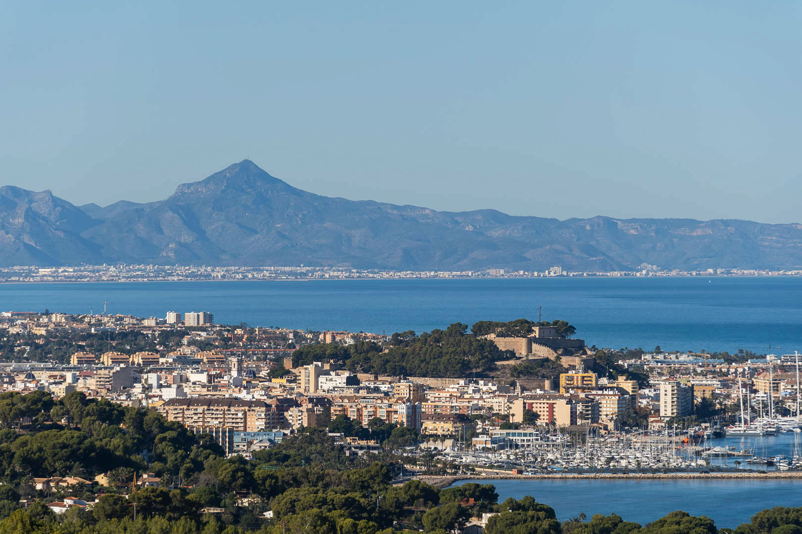 Panoramablick auf Dénia mit der historischen Burg, dem Yachthafen und der Mittelmeerküste vor der Kulisse der fernen Berge.