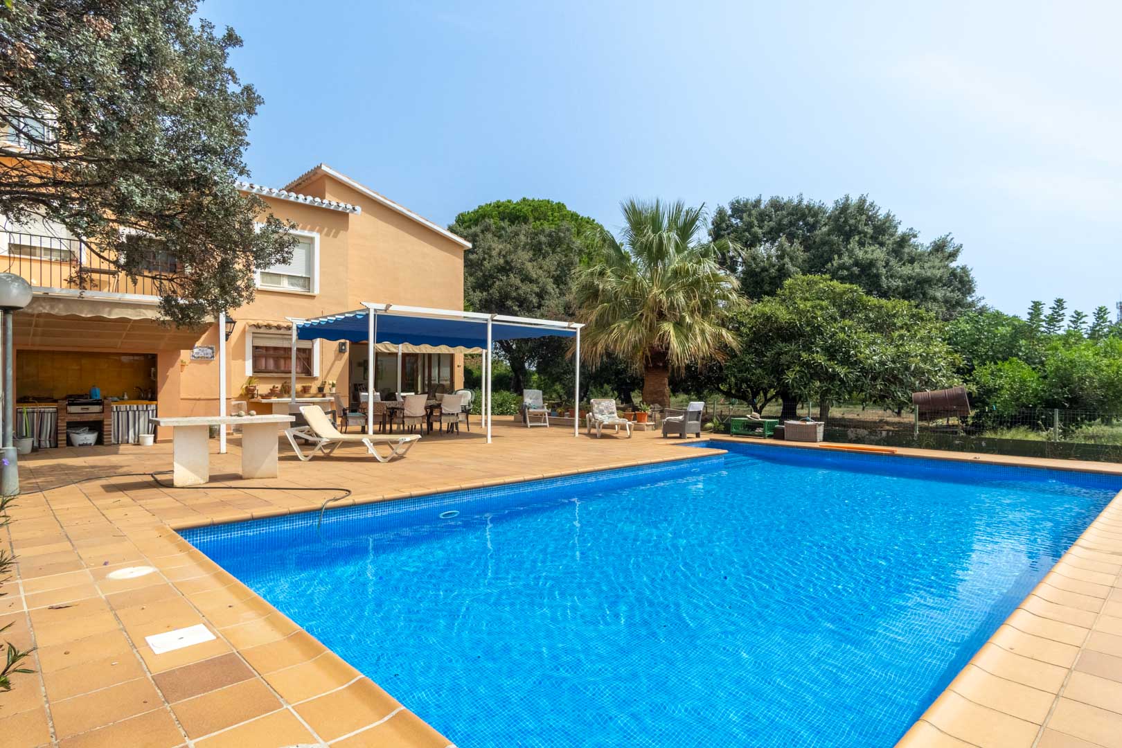 Rechteckiger Pool mit blauen Mosaikfliesen, umgeben von einer weitläufigen Terrasse, Außenküche und einem durch eine blaue Markise geschützten Essbereich.