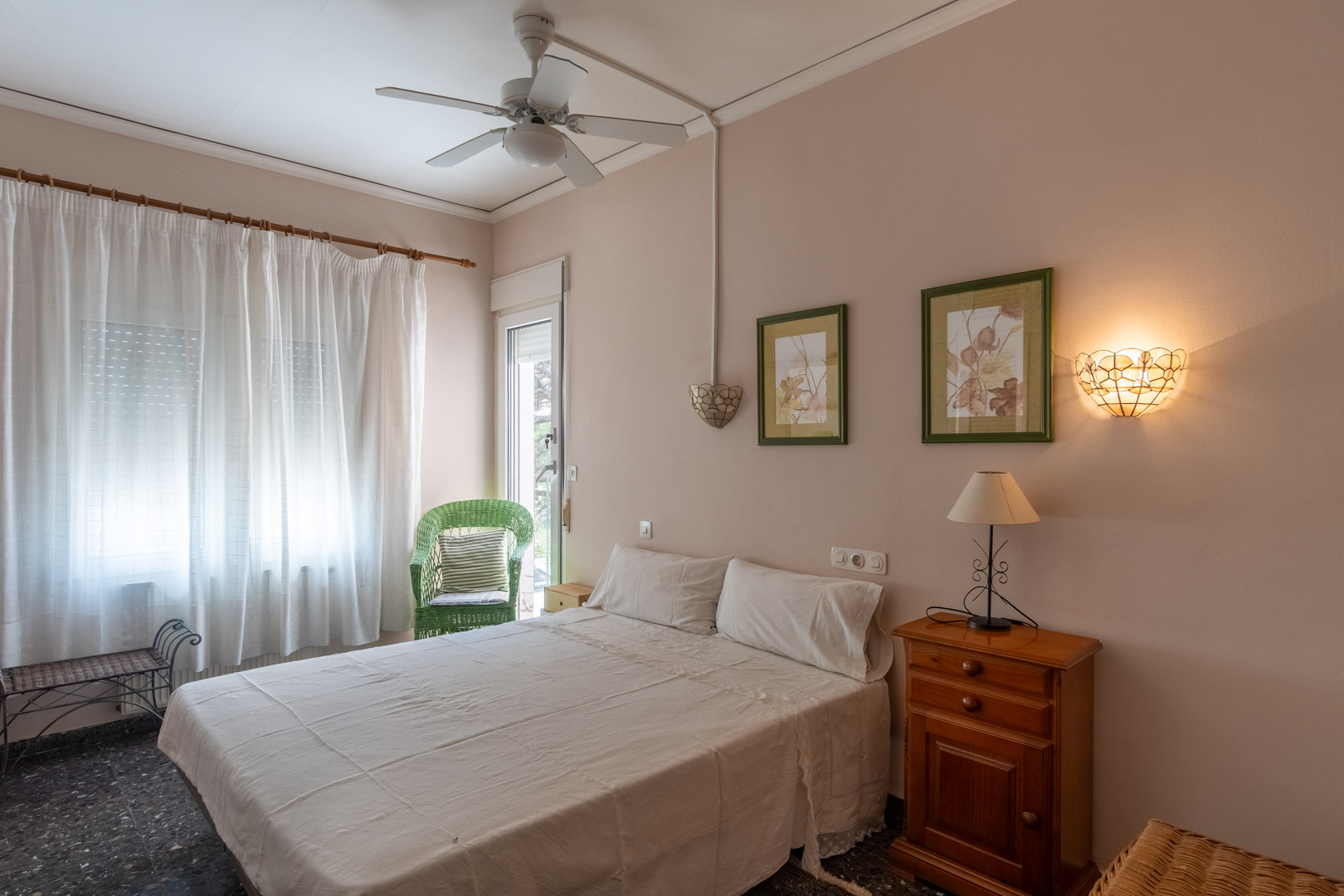 Schlafzimmer mit Doppelbett, Deckenventilator und direktem Zugang ins Freie. Mit Holzmöbeln, Terrazzoboden und einem dekorativen Korbsessel am Fenster.