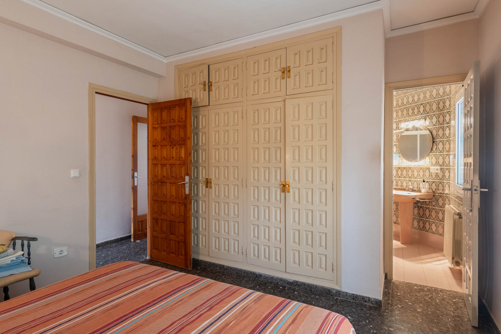 Schlafzimmer mit großem Einbauschrank mit Kassettenfronten und direktem Zugang zum En-Suite-Badezimmer mit geometrischen Fliesen und Tageslicht.