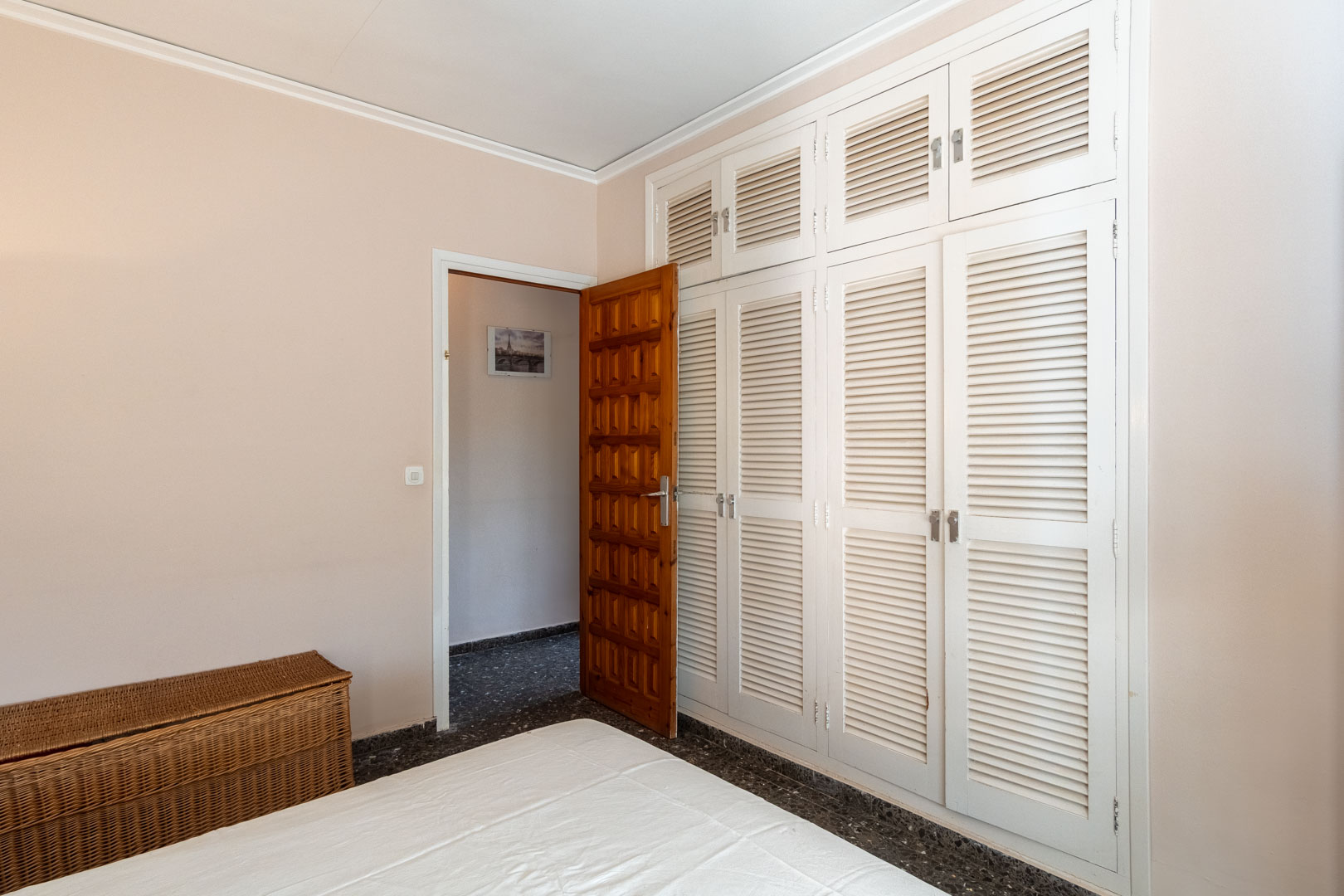 Schlafzimmer mit großem Einbauschrank mit weißen Lamellentüren, dunklem Terrazzoboden und einer massiven, verzierten Holztür zum Flur.