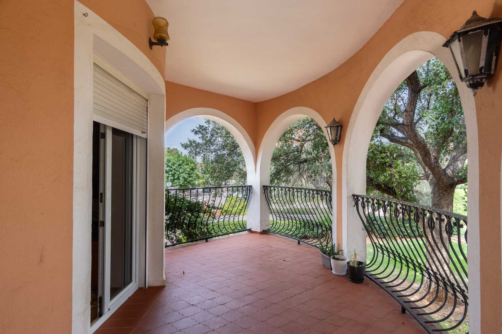 Überdachte Terrasse mit Rundbögen, geschwungenem Schmiedeeisengeländer und Terrakottaboden mit Blick auf den mediterranen Garten.
