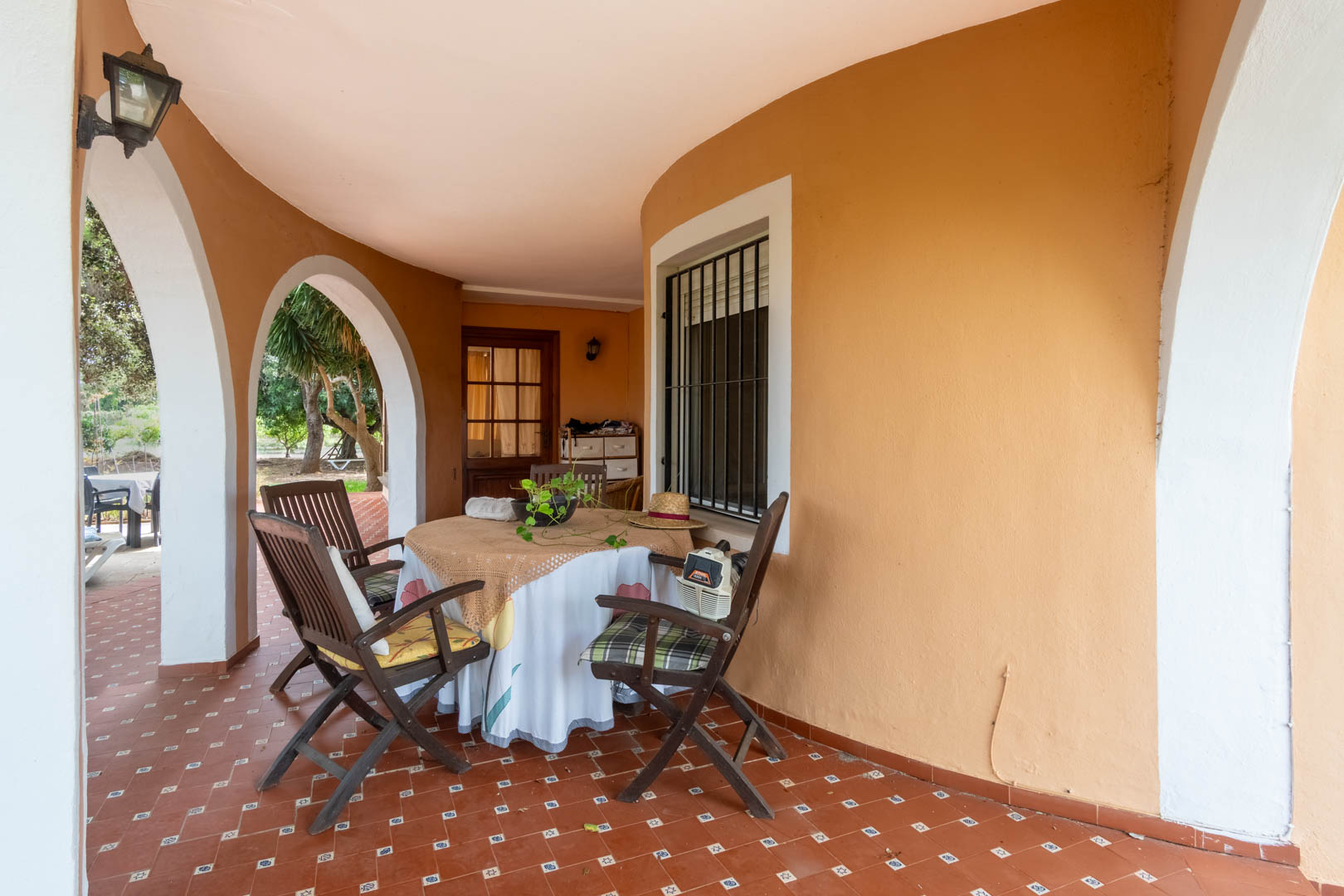 Überdachte Veranda mit weißen Rundbögen und rustikalem Fliesenboden. Ein schattiger Essbereich im Freien mit direktem Blick auf den mediterranen Garten.