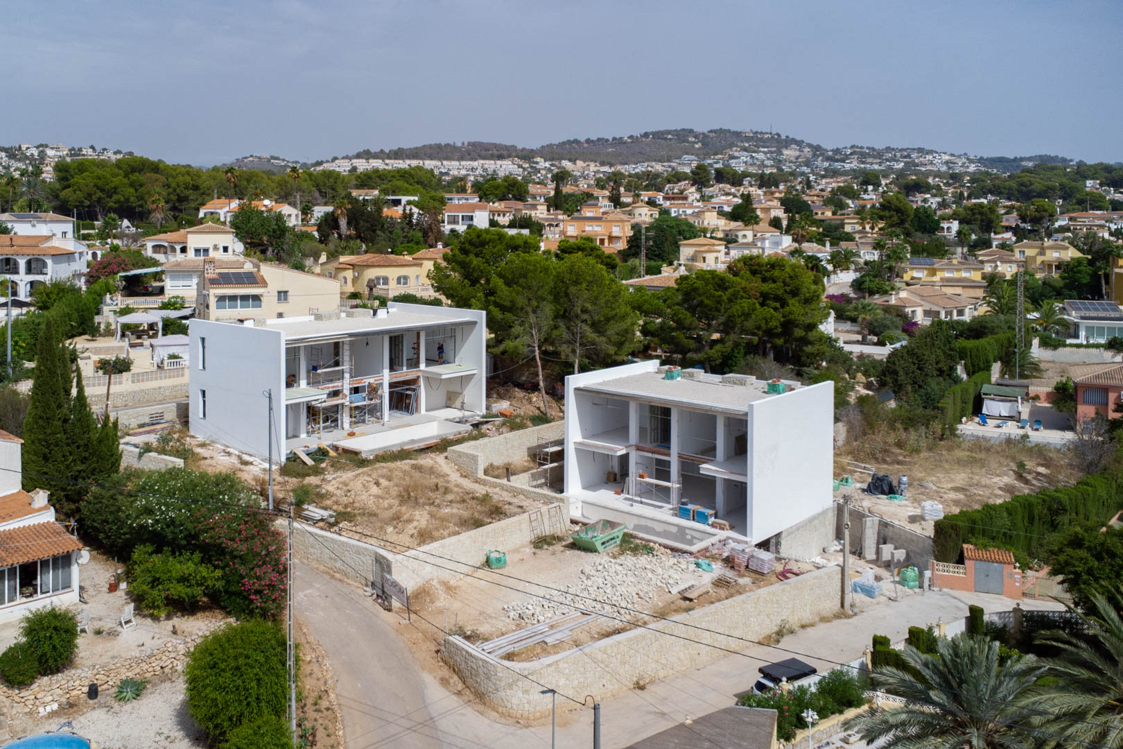 Luftaufnahme der Bauphase von zwei modernen Villen in Calpe, die die weißen Strukturen, Natursteinmauern und die Nivellierung der Grundstücke zeigt.