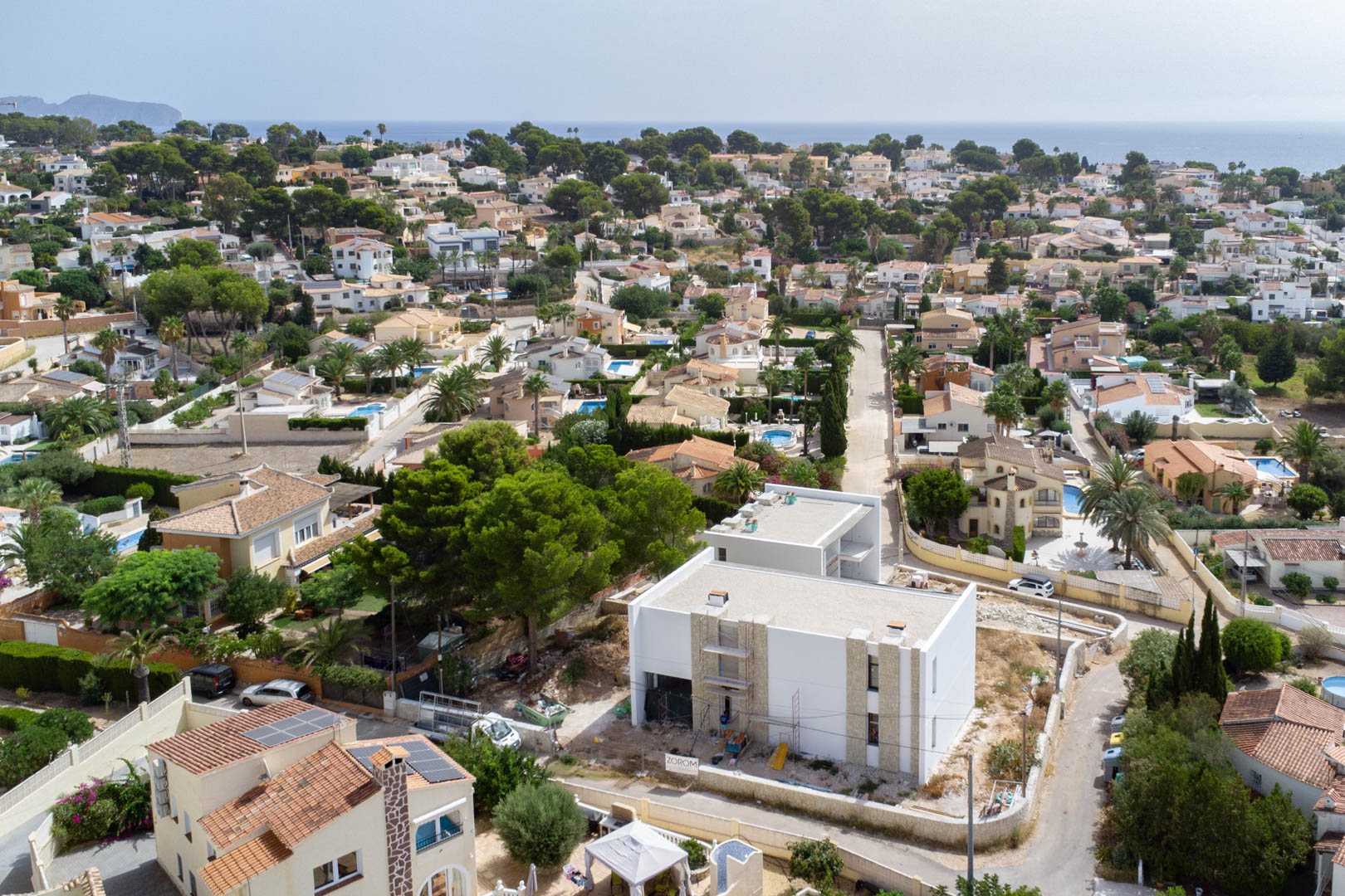 Luftaufnahme der Villa im Bau mit weißer Fassade und Natursteinelementen, eingebettet in ein Wohnviertel mit Blick auf das Mittelmeer am Horizont.