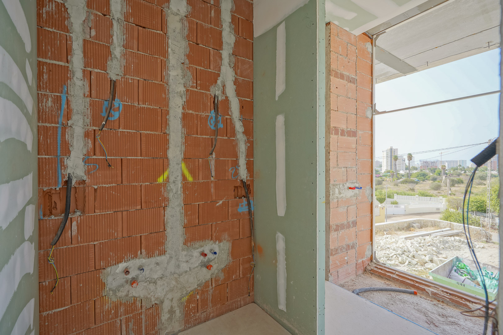 Innenansicht des Rohbaus mit Ziegelmauerwerk, verlegten Elektroleitungen und Sanitäranschlüssen neben einer bodentiefen Fensteröffnung.