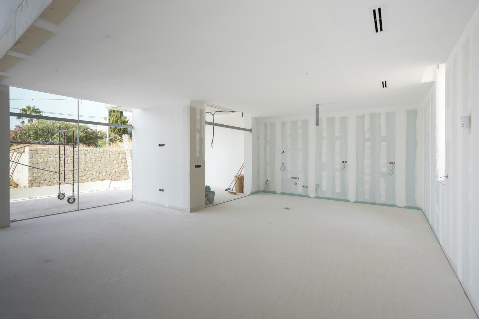 Innenansicht des Wohnbereichs im Rohbau mit integrierten Deckenauslässen für die Klimaanlage und großen Fensterfronten zur Terrasse.