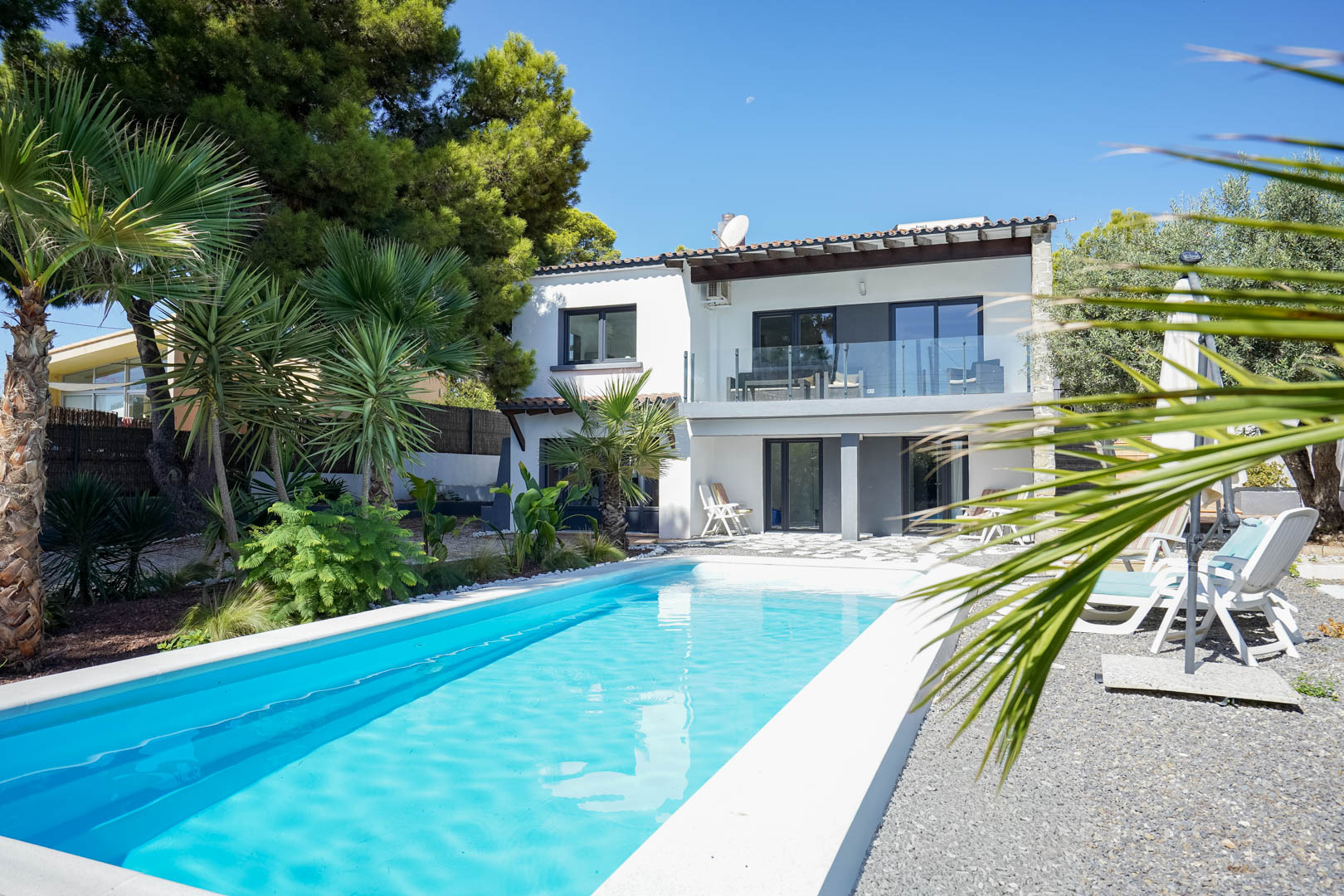 Außenansicht der Villa mit rechteckigem Pool, mediterranem Garten mit Palmen und zweistöckiger weißer Fassade mit Glasbalkonen.
