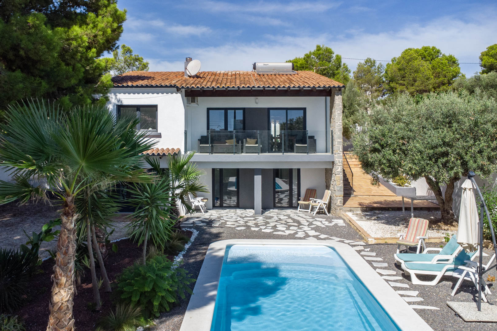 Außenansicht der Villa mit rechteckigem Pool, mediterranem Garten und weißer Fassade. Mit Glasbalkonen und einer Solarthermieanlage auf dem Ziegeldach.