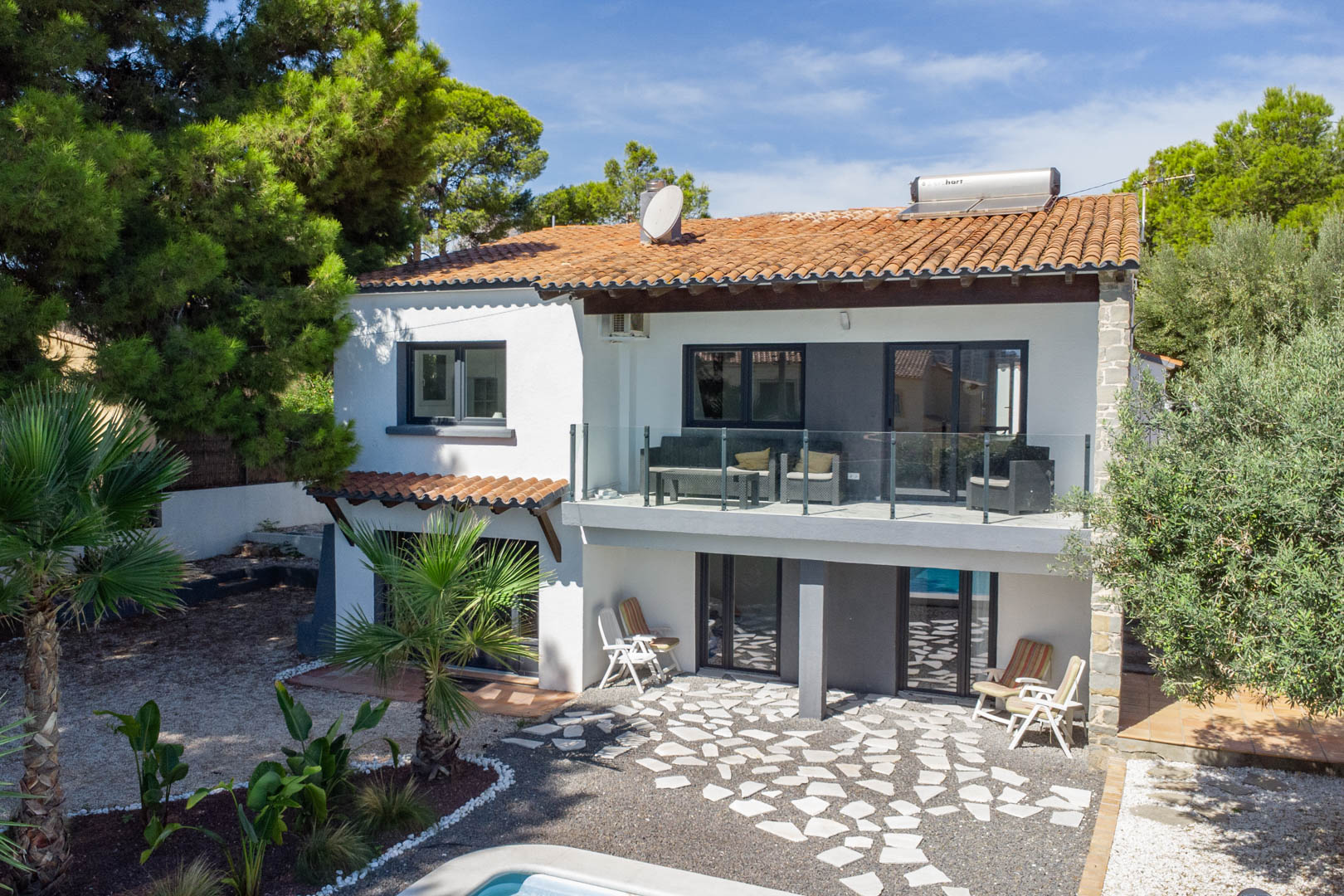 Außenansicht der zweistöckigen Villa mit Glasbalkon, Solarthermie auf dem Ziegeldach und einem gepflasterten Außenbereich im Erdgeschoss.