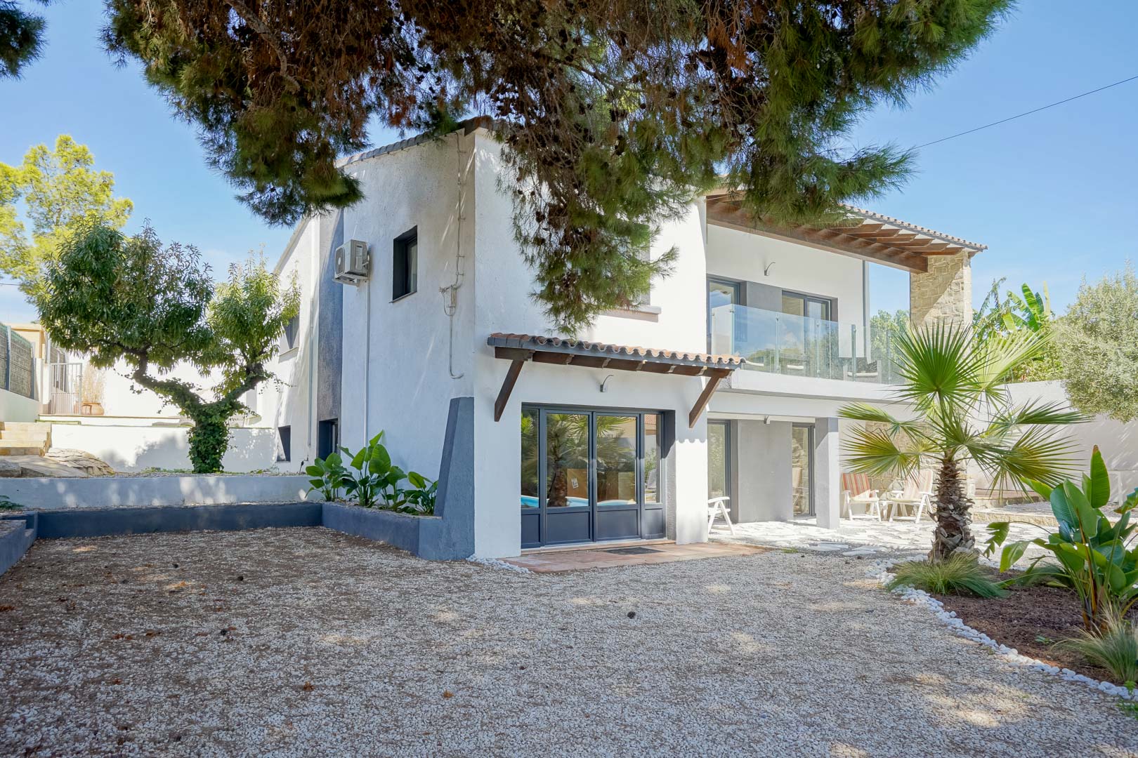 Außenfassade der Villa mit grauen Fensterrahmen, Glasgeländer-Terrassen und einem mediterranen Garten mit Kiesflächen und Palmen.