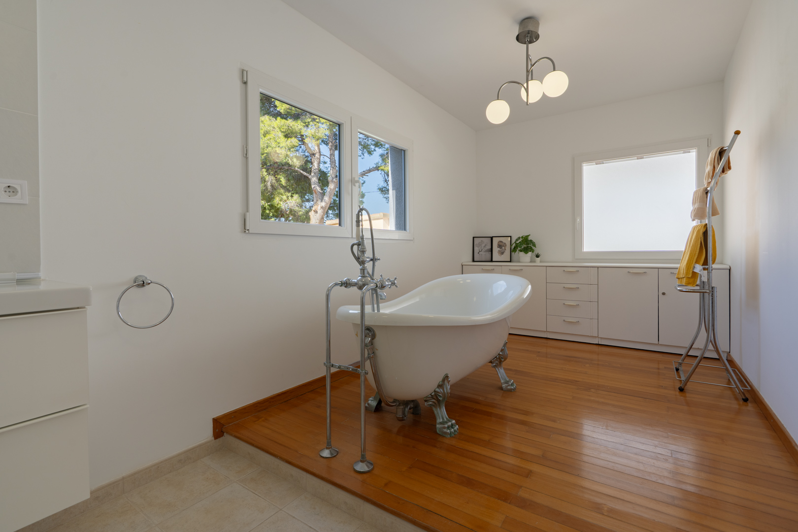 Badezimmer mit freistehender Klauenfuß-Badewanne auf einem Holzpodest, weißen Einbauschränken und großen Fenstern für reichlich Tageslicht.